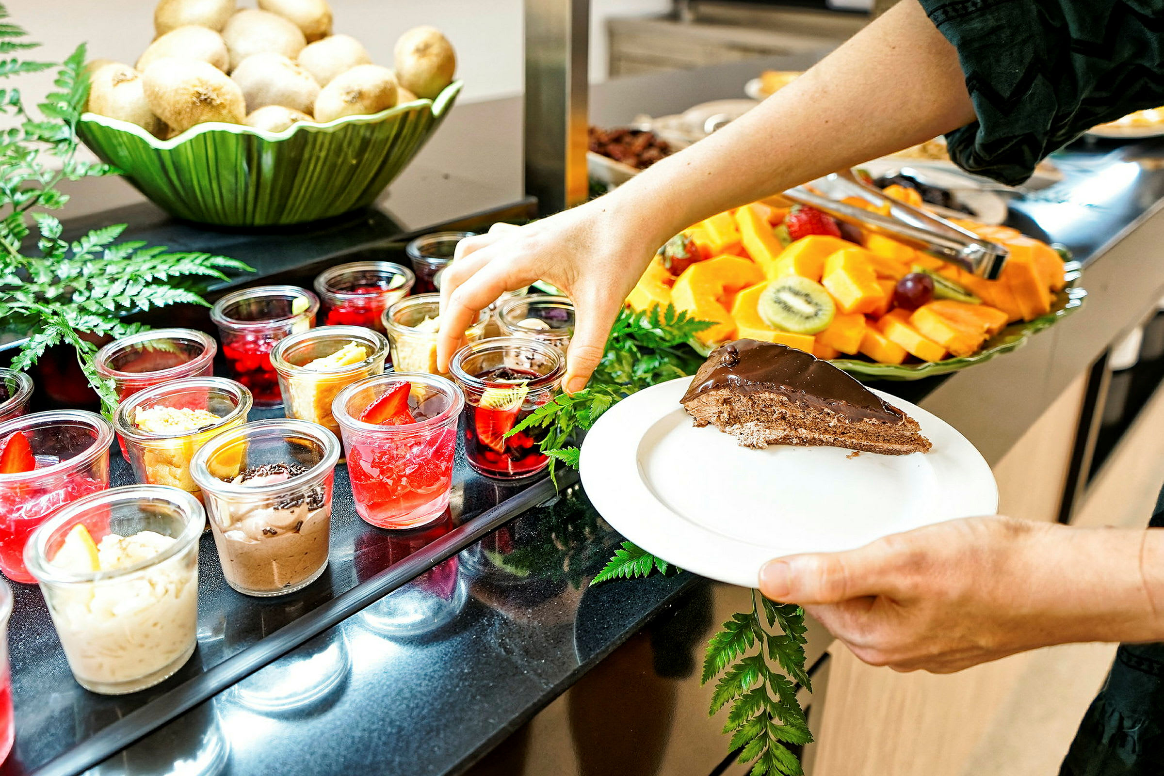 En person tar en dessert fra en buffé med et utvalg av små desserter i glass, frukter og et stykke sjokoladekake på en tallerken. (AI-generert tekst)
