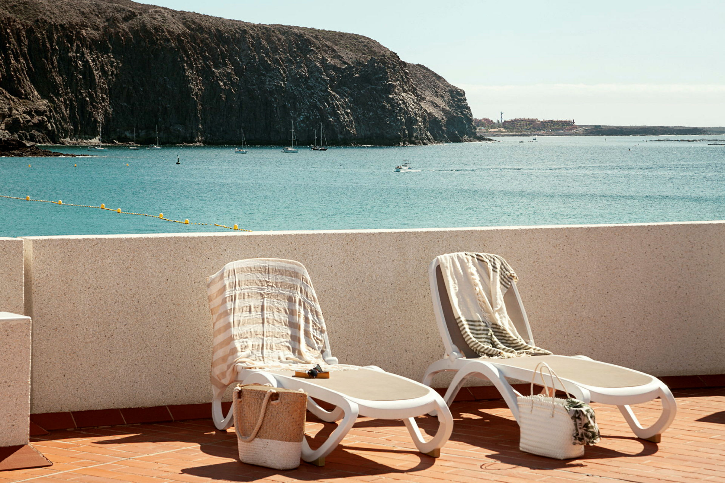 To solsenger med håndklær og kurver, plassert på en terrasse med utsikt over havet og en klipperik kystlinje. (AI-generert tekst)