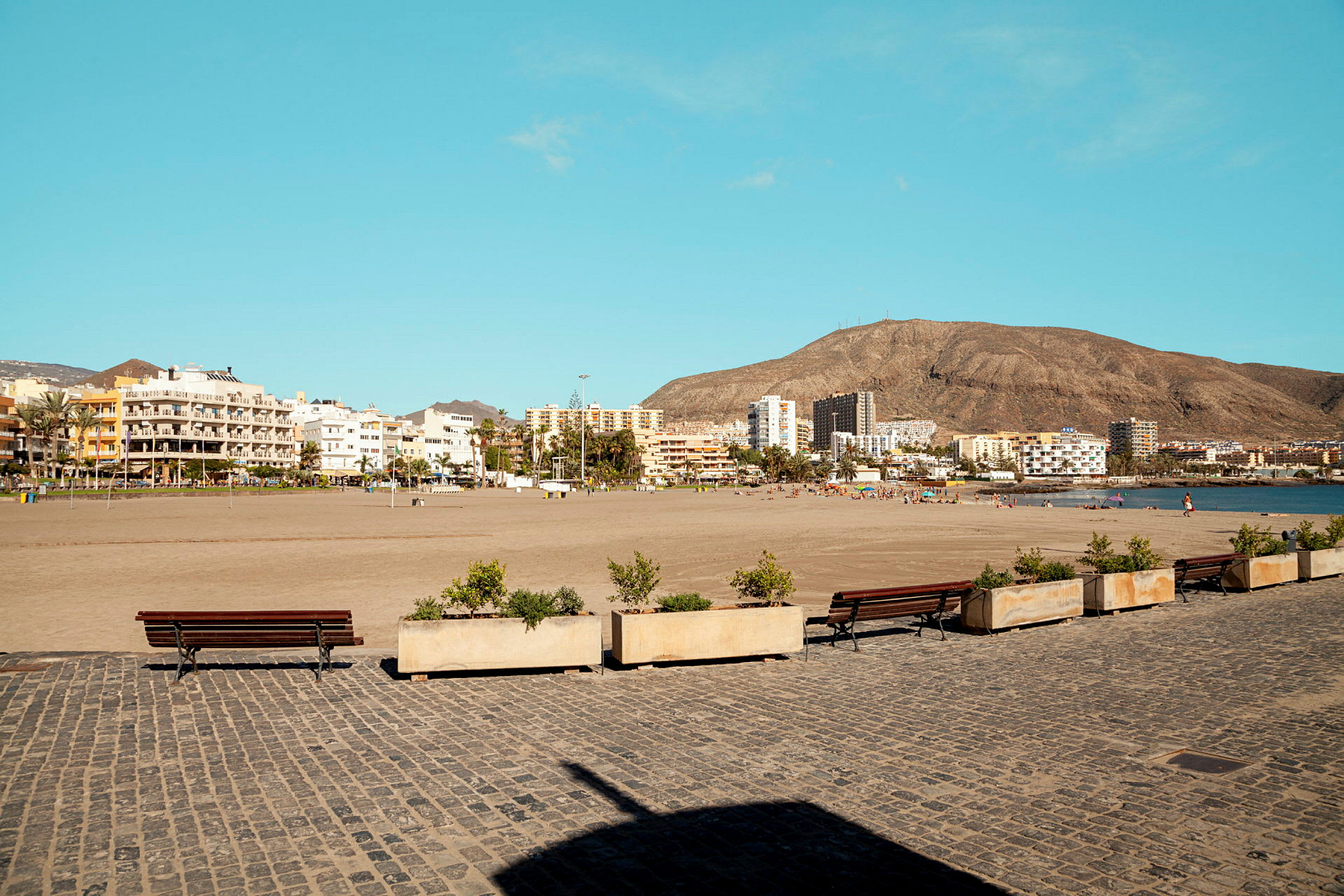 En sandstrand med mennesker i bakgrunnen, byen sees lenger unna ved foten av en fjellkjede. I forgrunnen er det en steinbelagt gangvei med benker og blomsterkasser. (AI-generert tekst)