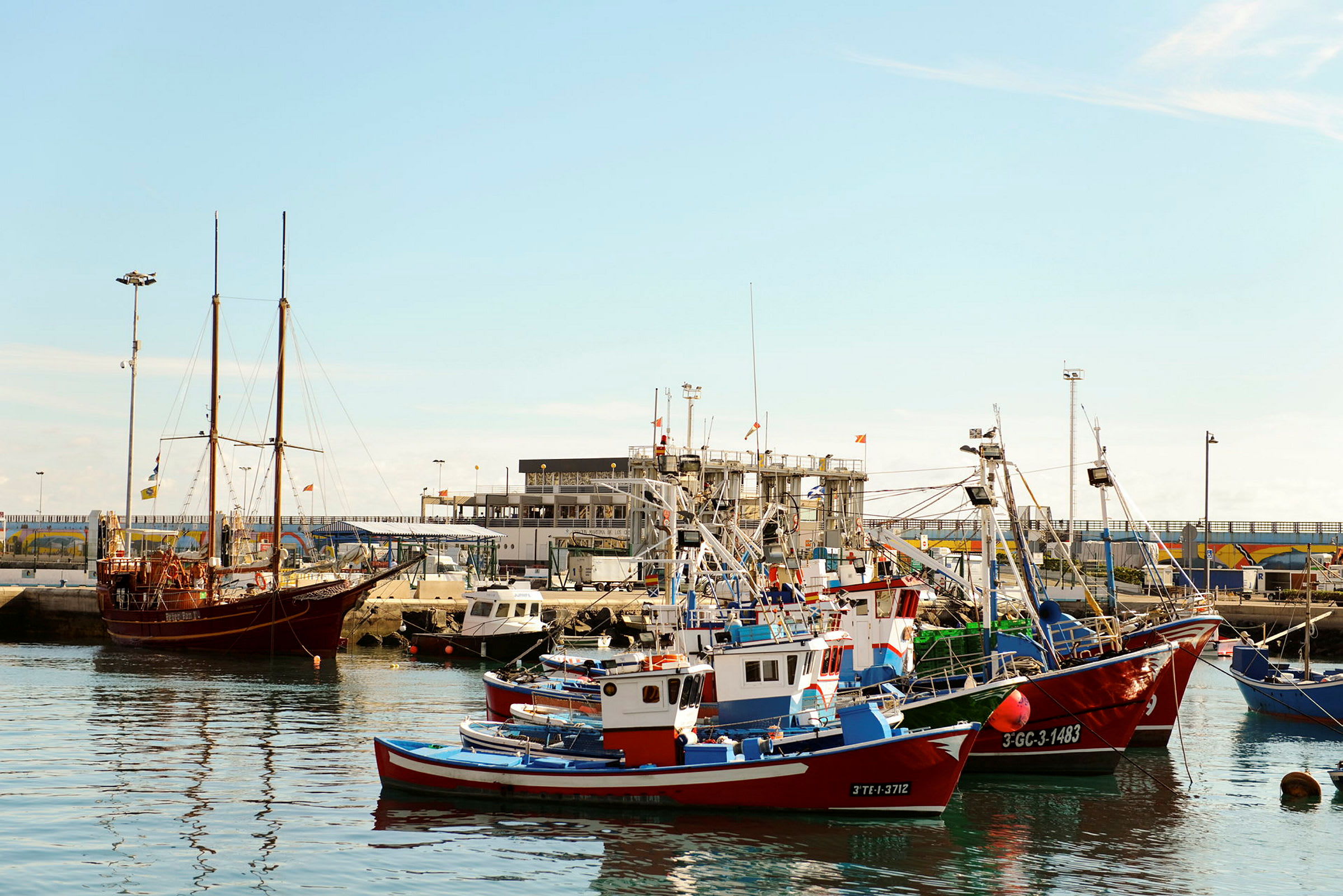 Bildet viser en havn med flere fiskebåter og en større seilbåt ved kaien. (AI-generert tekst)