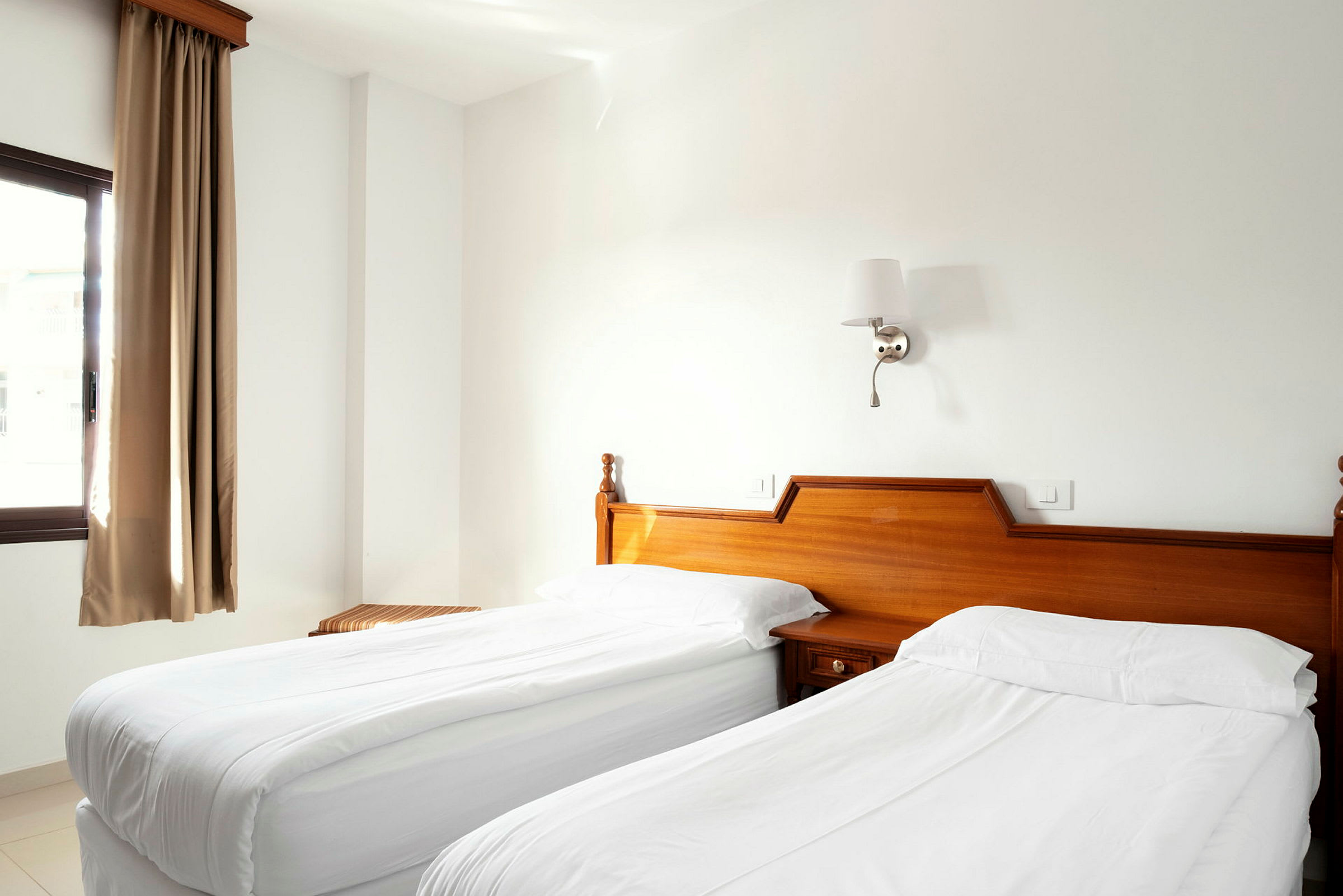 Et hotellrom med to enkeltsenger dekket med hvitt sengetøy, tremøbler og et vindu med beige gardiner. (AI-generert tekst)
