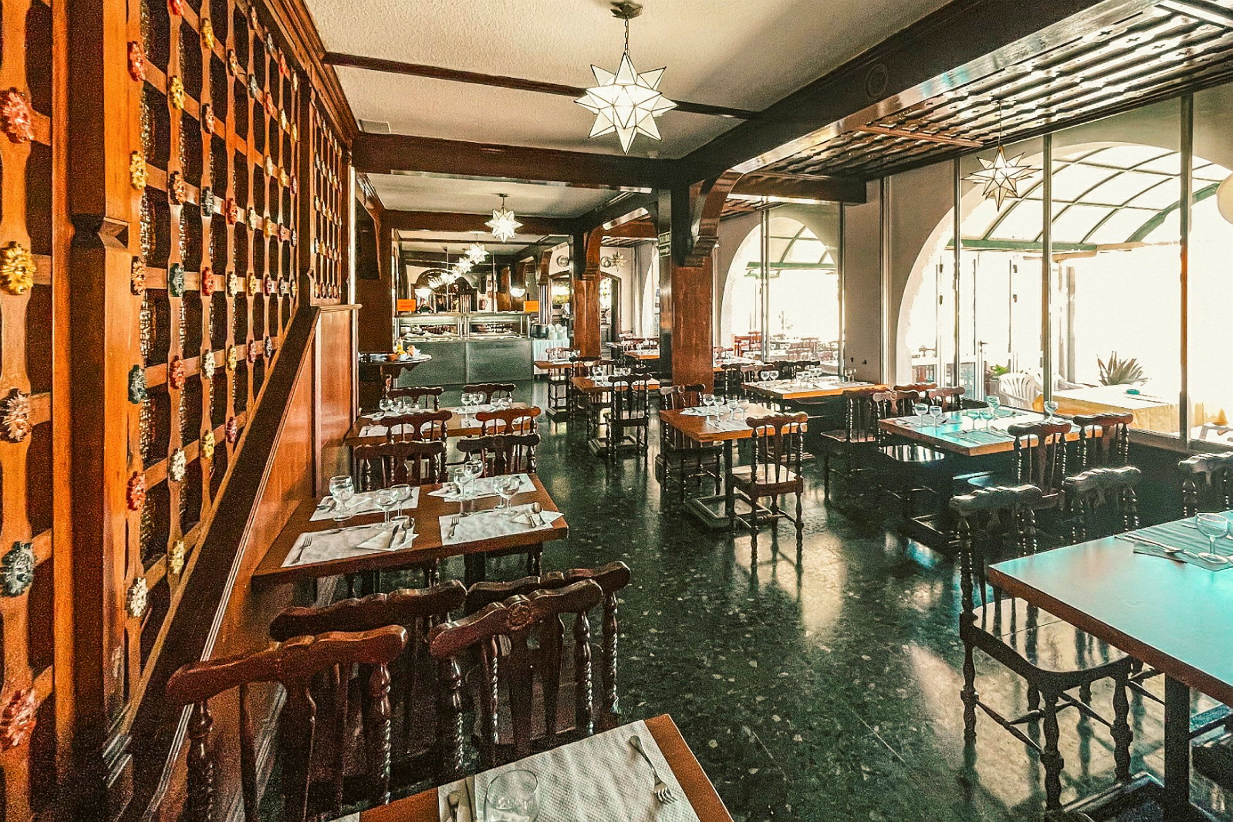 En restaurant med mørke tremøbler og dekorerte taklamper. Bordene er dekket med tallerkener og glass. (AI-generert tekst)
