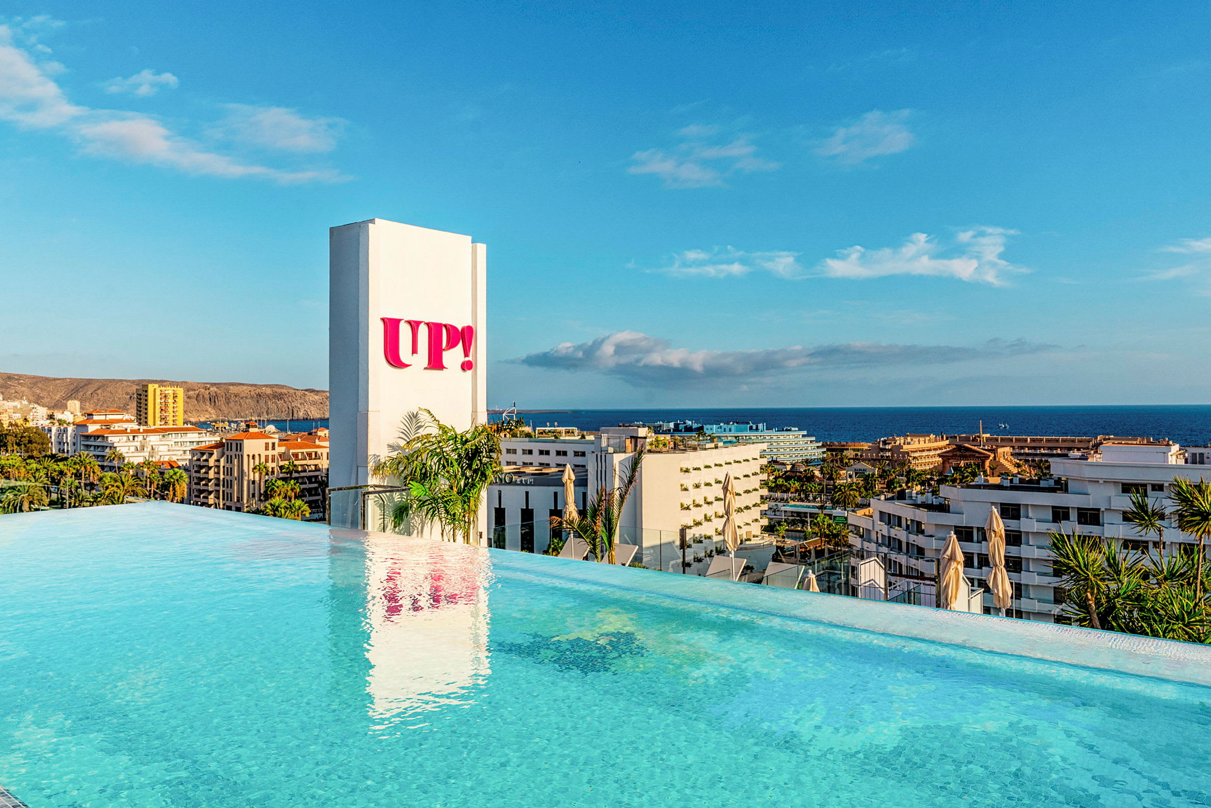 Utsikt over et infinitybasseng med havet og bygninger i bakgrunnen. Et skilt med teksten "UP!" er synlig ved siden av. (AI-generert tekst)