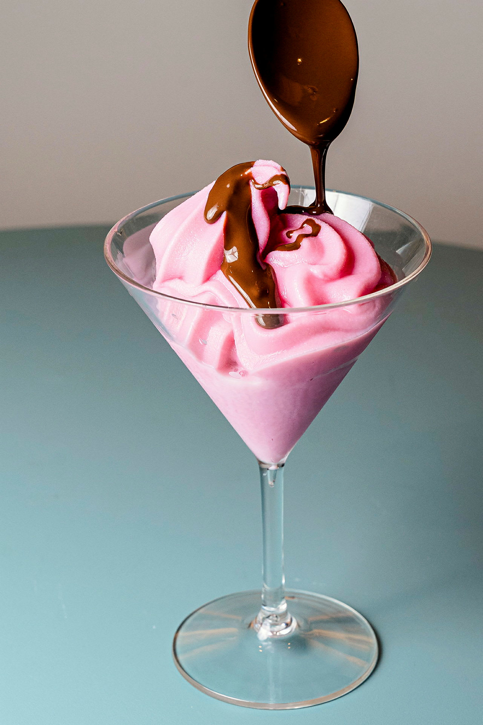 Et glassbeger fylt med rosa iskrem og dekorert med sjokoladesaus som helles fra en skje. (AI-generert tekst)