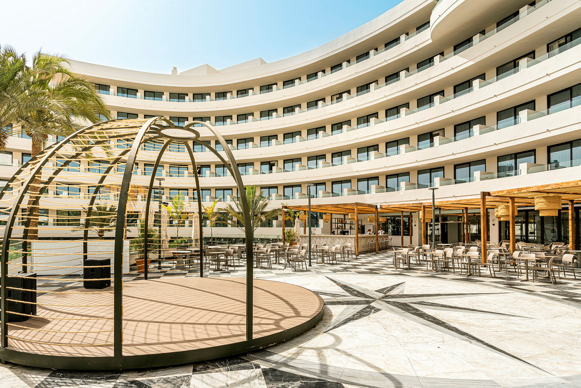 Bilde av en moderne fleretasjes hotellbygning med balkonger rundt en stor gårdsplass med sitteplasser, palmer og en robust trestruktur. (AI-generert tekst)