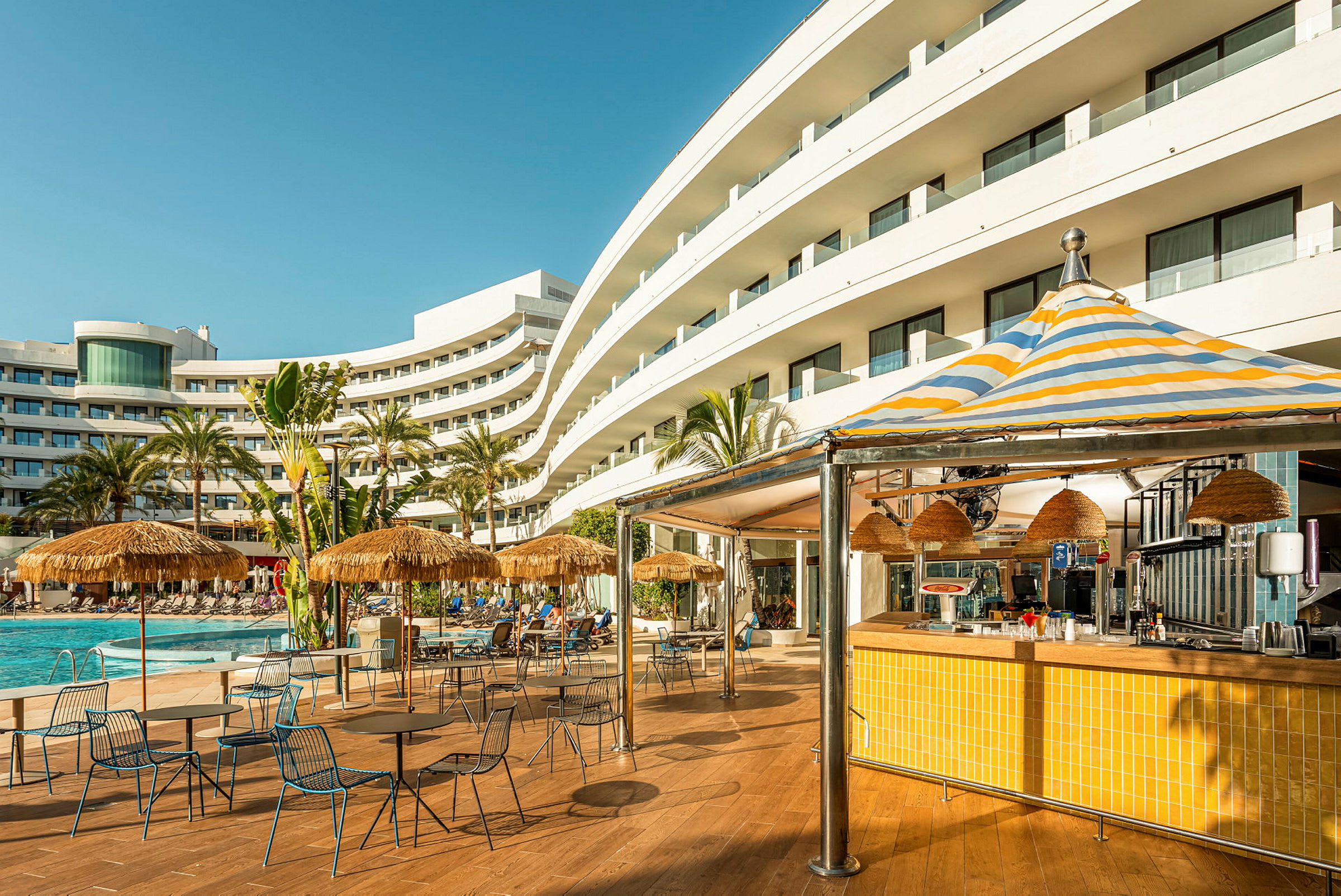 Et hotell sett utenfra med bassengområde, parasoller og en serveringsbar. (AI-generert tekst)