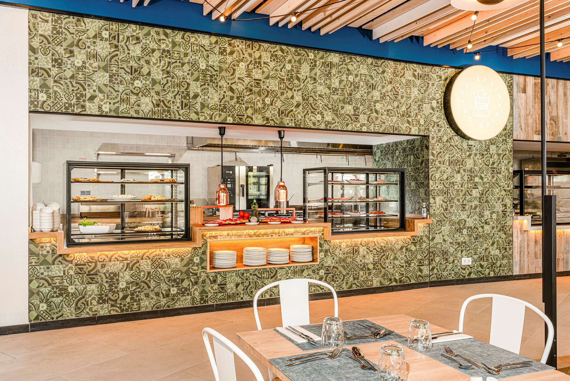 En opplyst buffet med forskjellige matretter og kjøkkenutstyr bak glasskjermer i en restaurantmiljø. Bord med duker og glass sees i forgrunnen. (AI-generert tekst)