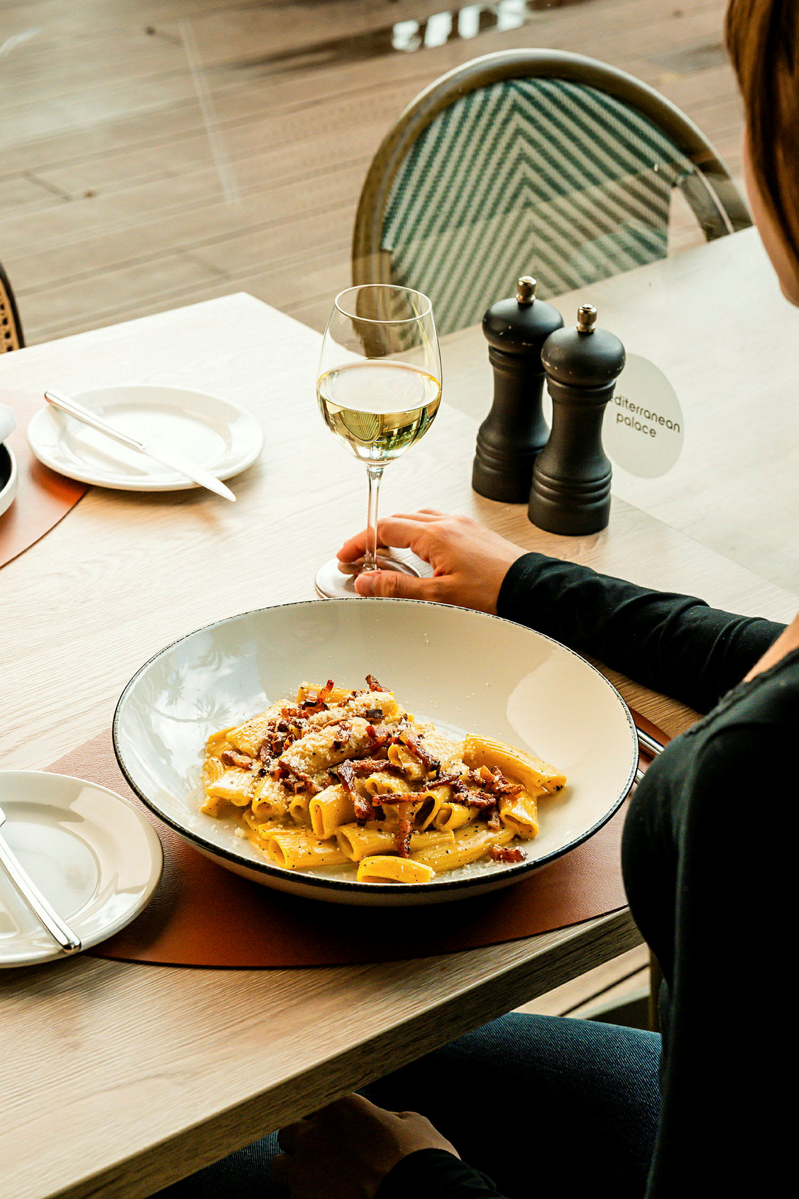 En tallerken pasta toppet med bacon og ost ved siden av et vinglass og et par krydderkverner. (AI-generert tekst)