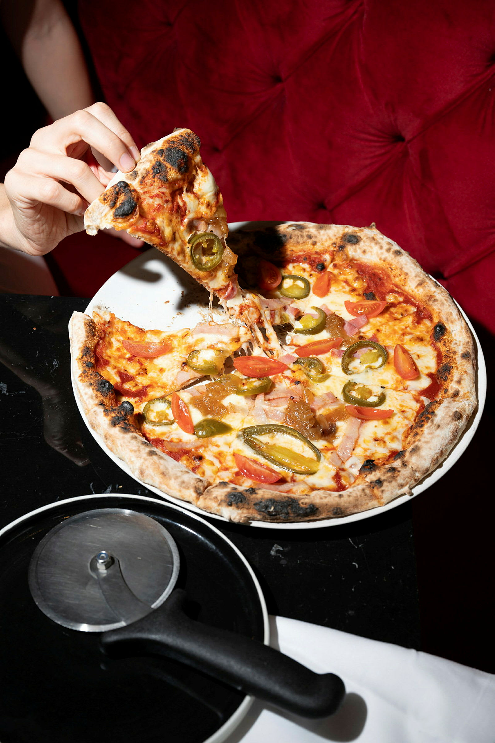 En hånd holder et pizzastykke med pålegg som jalapeño og tomat, og en pizzaskjærer ligger på bordet ved siden av en tallerken med resten av pizzaen. (AI-generert tekst)