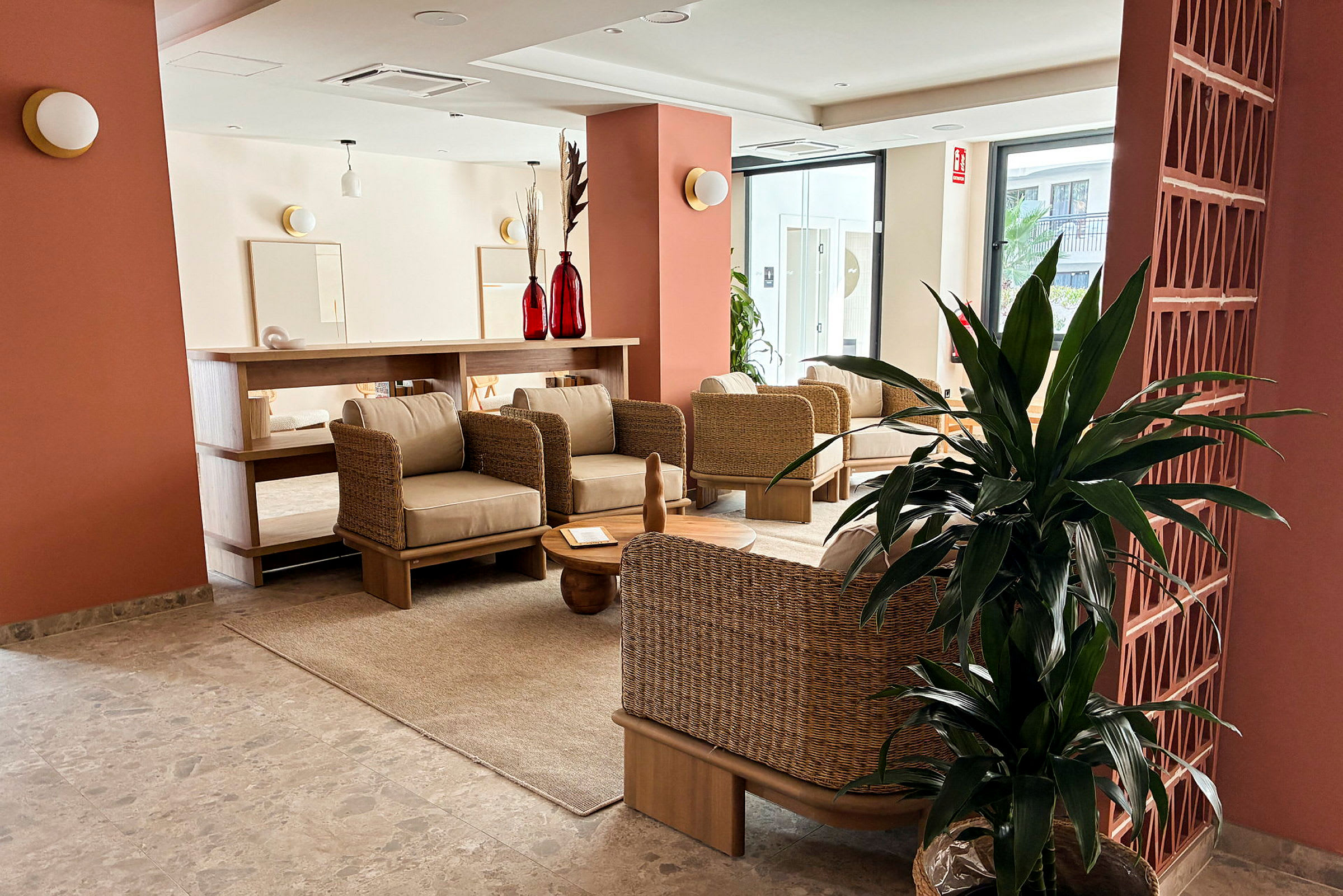 En lounge med rottingmøbler, et beige teppe og røde dekorative objekter på et bord. Planter og store vinduer ses i bakgrunnen. (AI-generert tekst)
