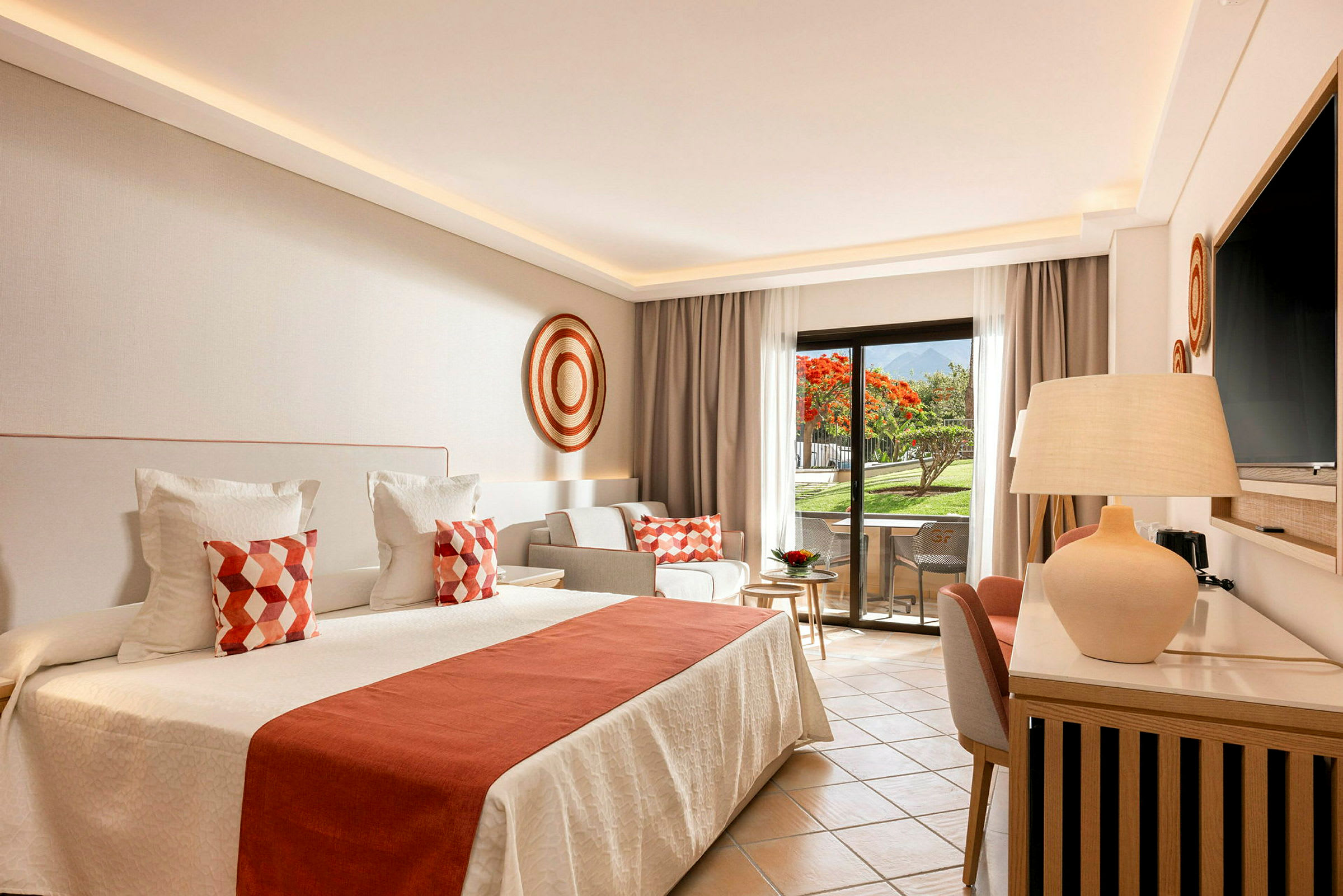Et lyst og elegant hotellrom med en stor seng, dekorerte puter og et privat uteseteområde med utsikt over en hage. (AI-generert tekst)