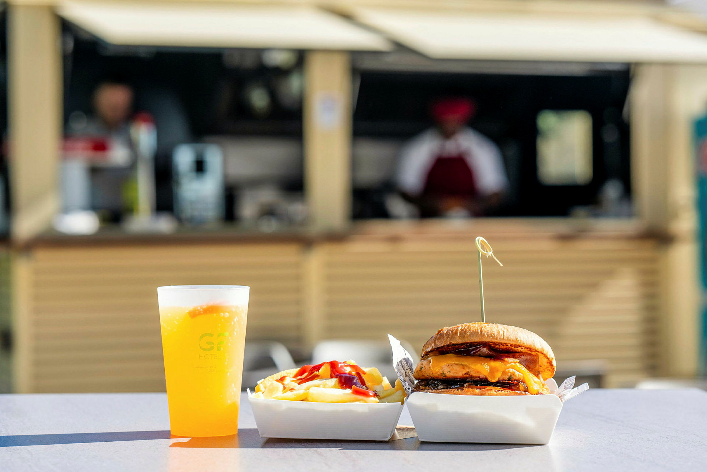 Mat som består av en hamburger, pommes frites og en drikke i en plastkopp på et bord utendørs, med bakgrunn av en serveringsbod. (AI-generert tekst)