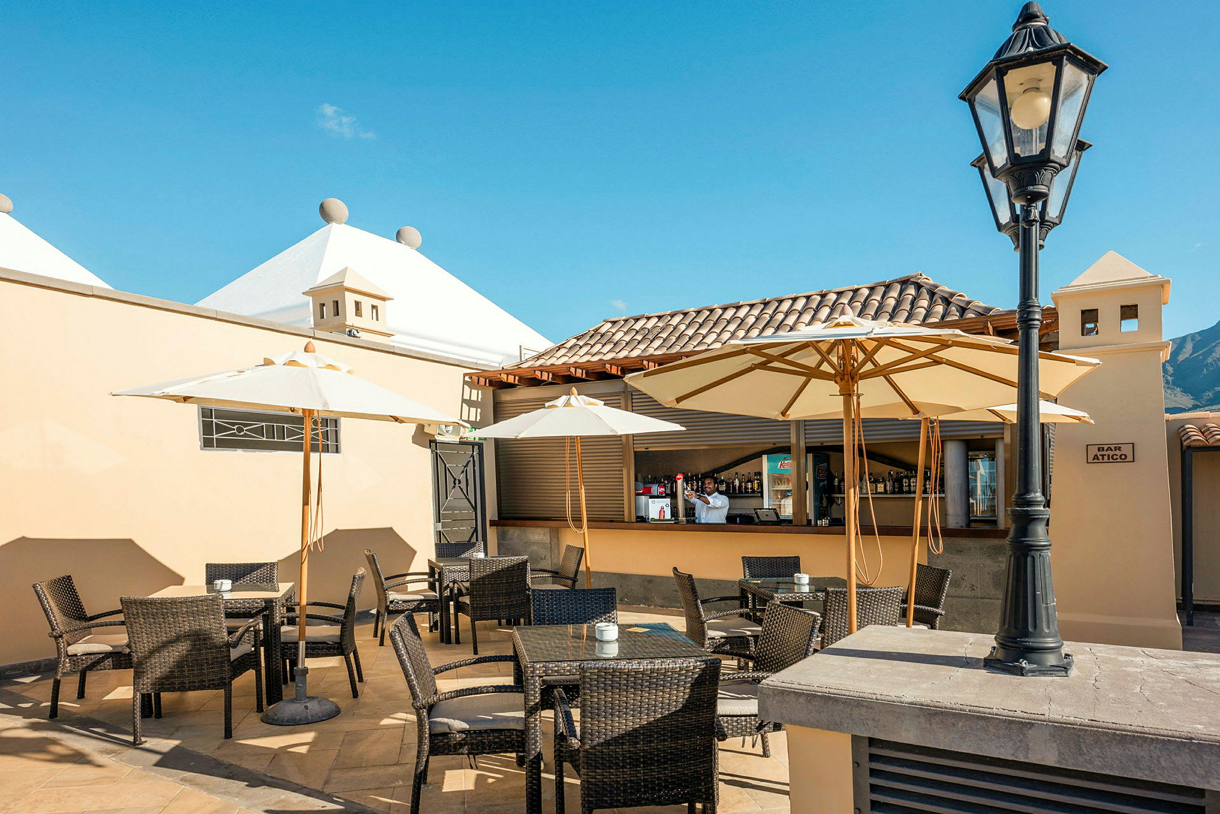 En utendørsrestaurant med bord, stoler og parasoller under en klarblå himmel. (AI-generert tekst)