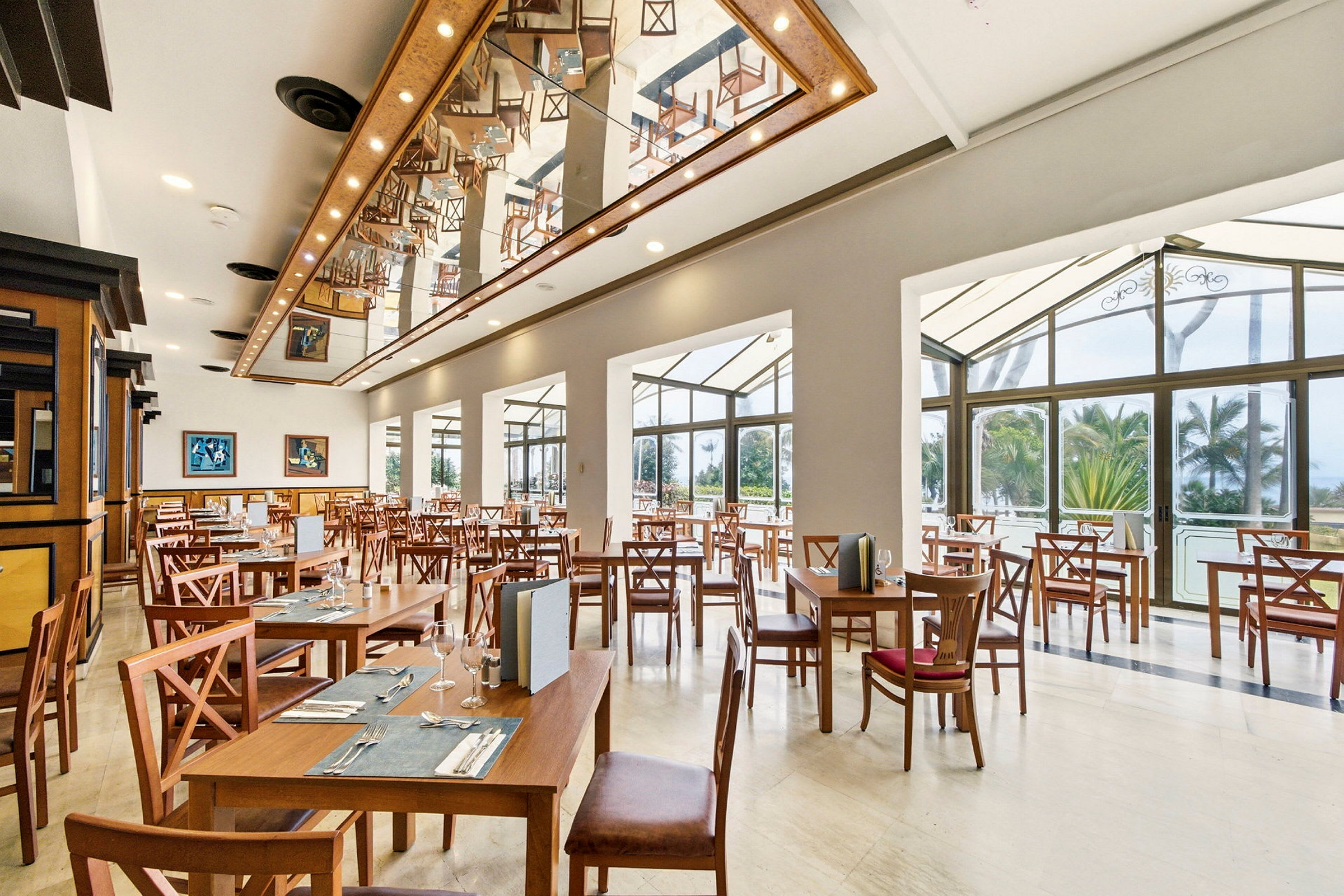En lys og romslig restaurant med trebord og stoler, takdekorasjoner og store vinduer med utsikt mot grønne omgivelser utenfor. (AI-generert tekst)
