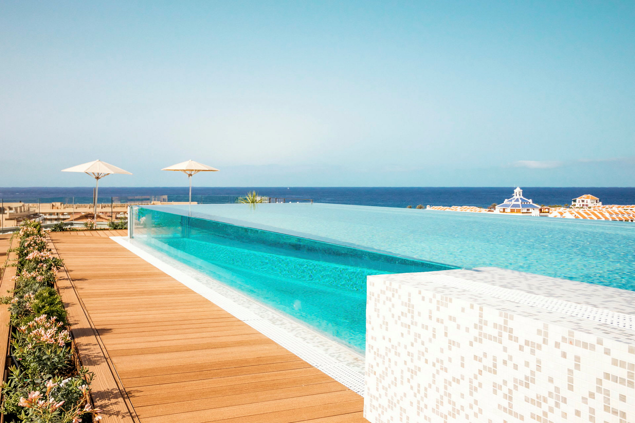 Et langt infinitybasseng med utsikt over havet og omgivelsene, omgitt av terrassebord og parasoller. (AI-generert tekst)