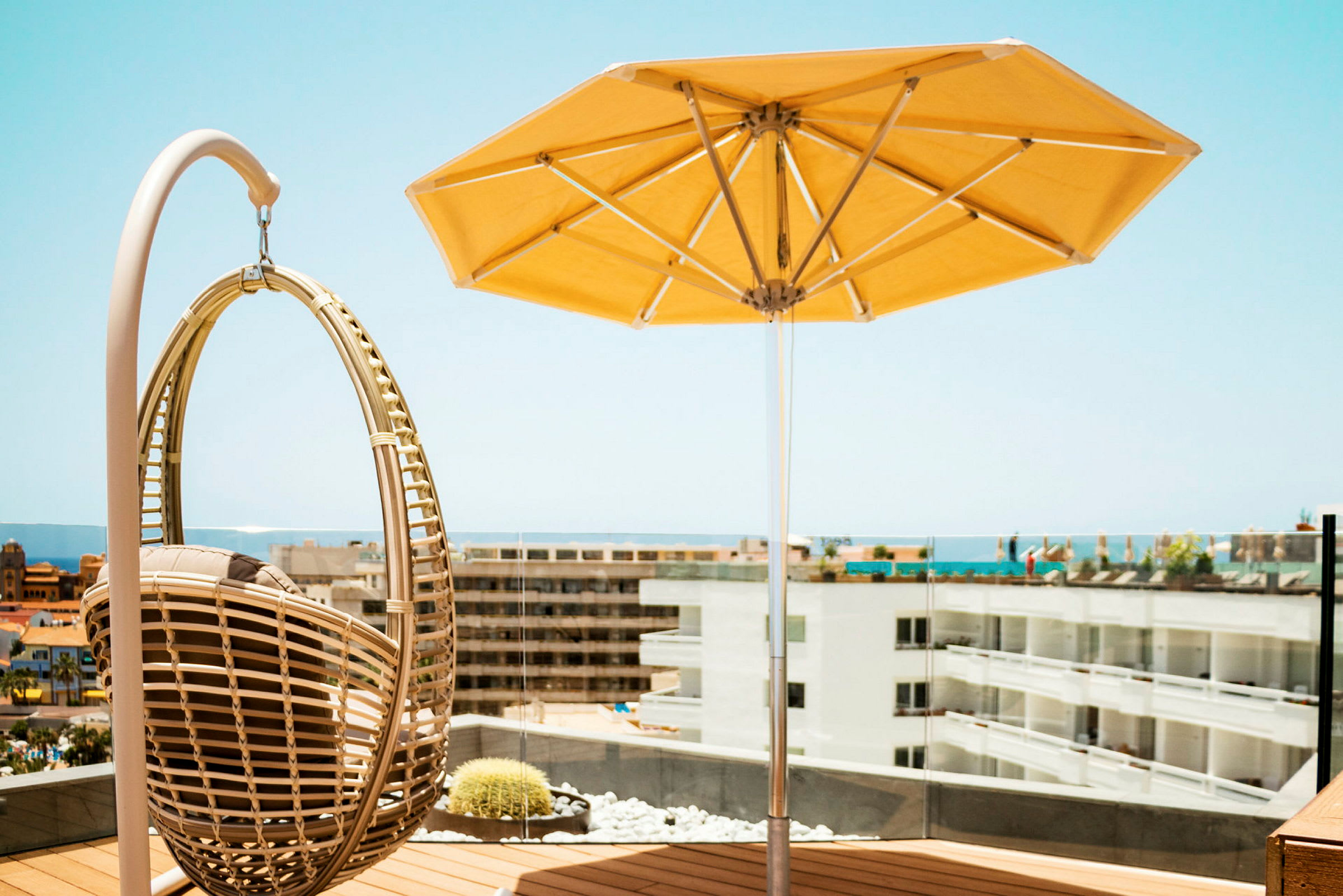 En hengende stol og en gul parasoll på en terrasse med utsikt over bygninger og havet. (AI-generert tekst)