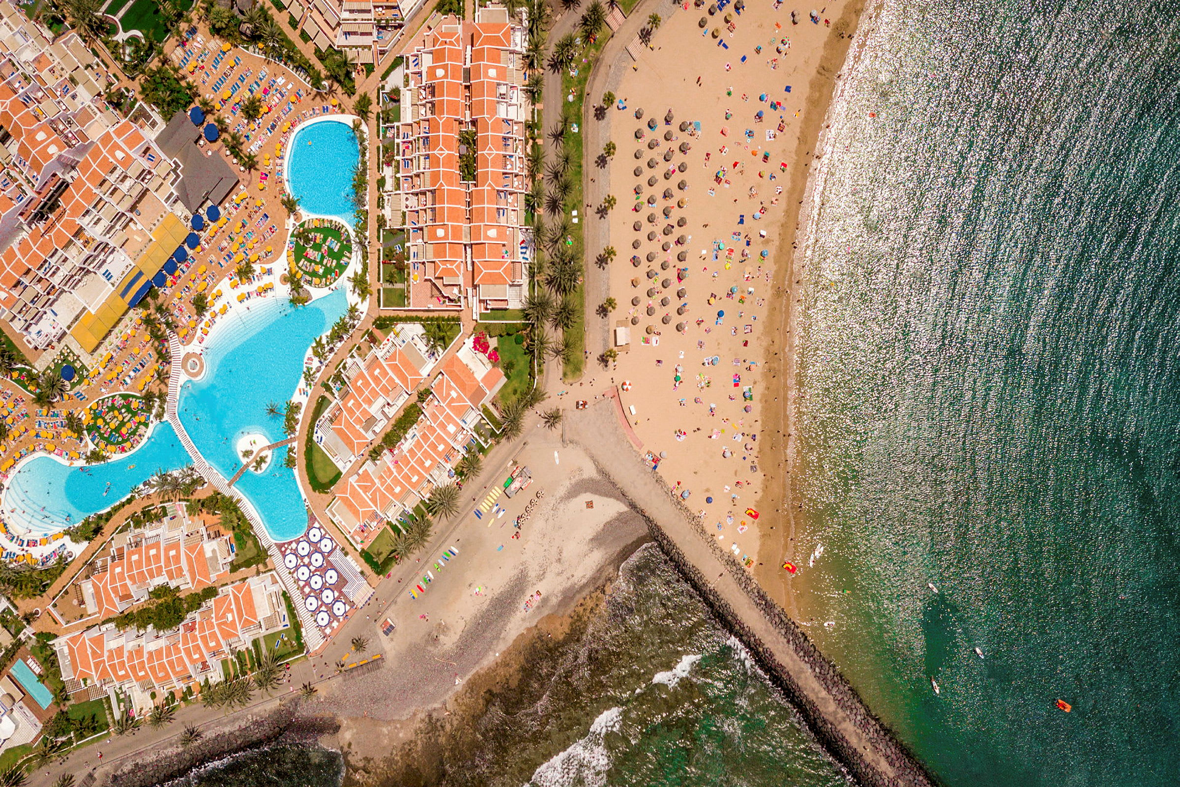 Luftfoto av en strand med solskjermer, en havsbukt og tilstøtende hotelldommer med bassenger. (AI-generert tekst)
