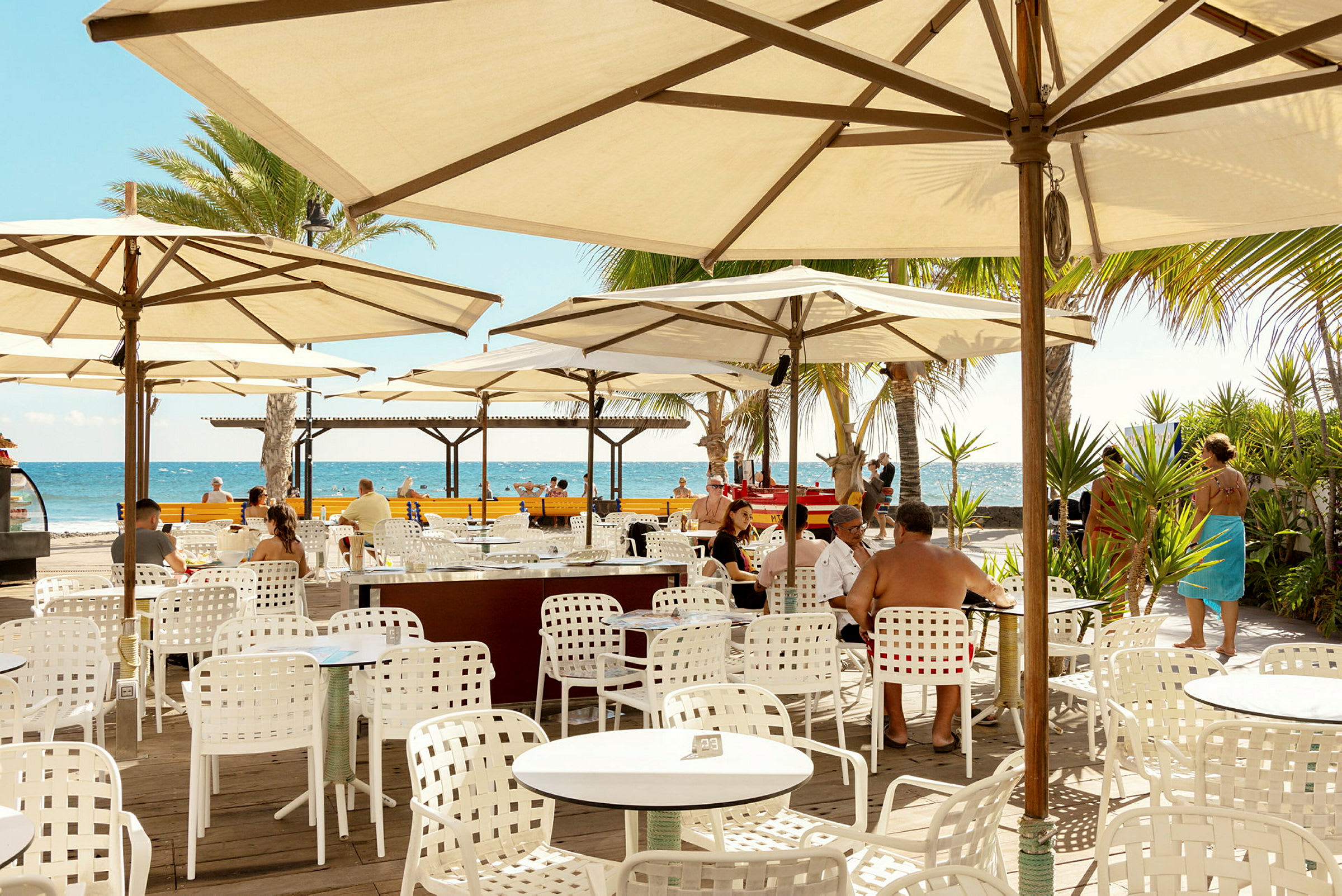 Utendørs restaurant med hvite bord og stoler under parasoller, utsikt mot havet. (AI-generert tekst)