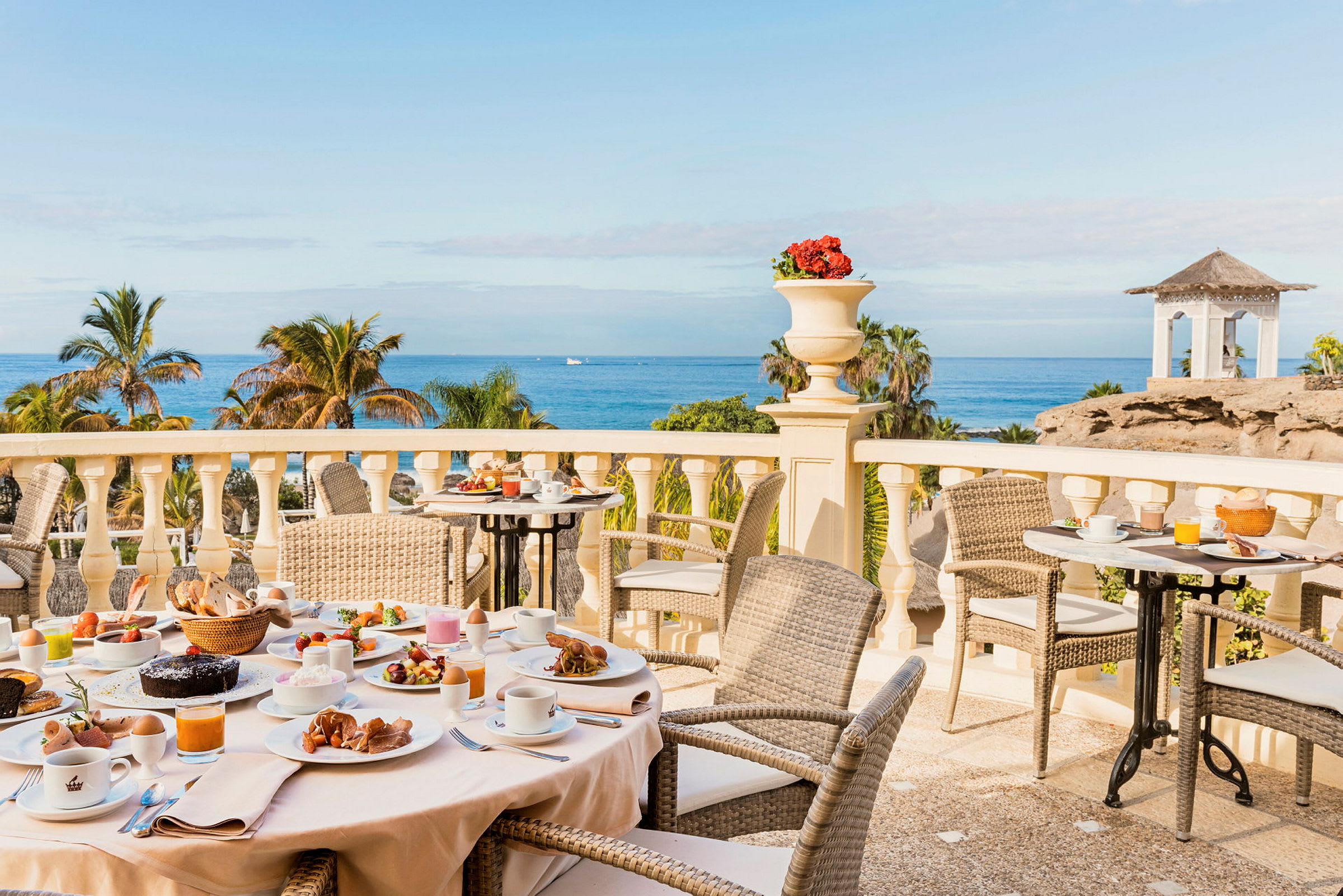 Frokostbord med forskjellige retter på en terrasse med utsikt over palmer og havet. (AI-generert tekst)