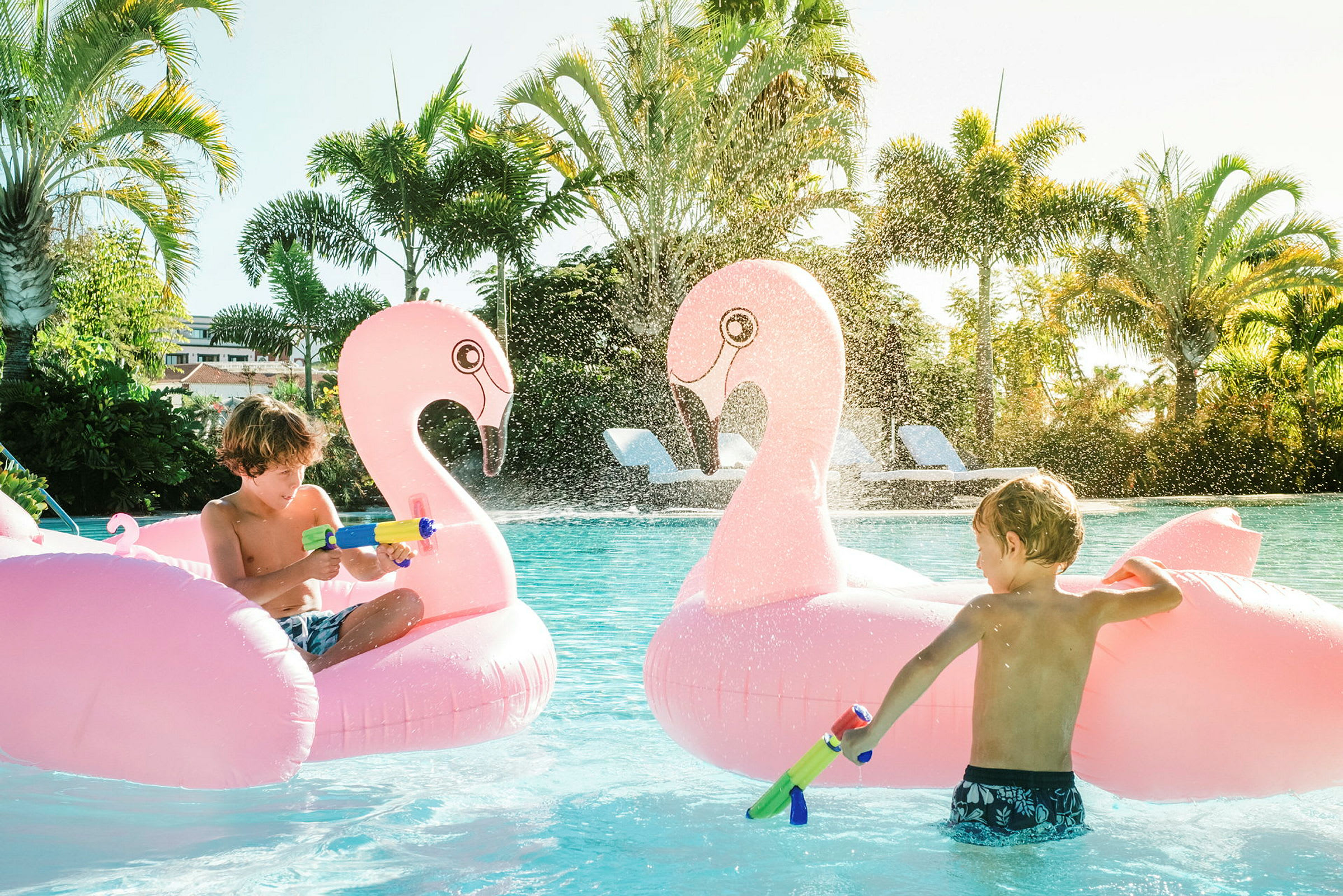 To oppblåsbare flamingo-figurer i et basseng med vannstråler og omgivelser med palmer. (AI-generert tekst)