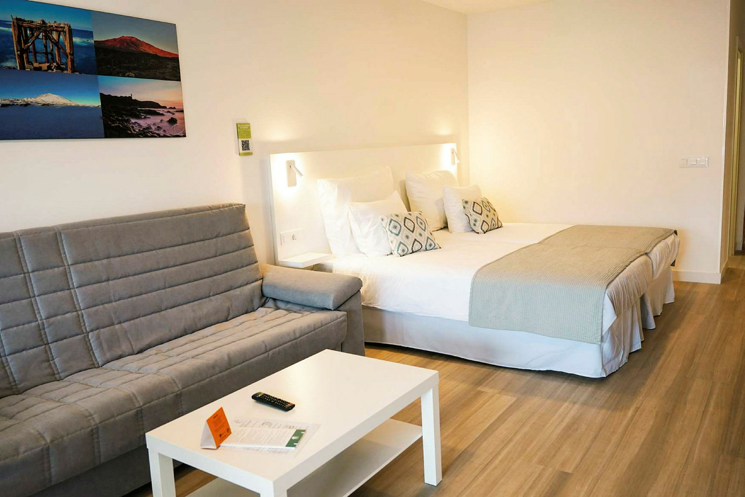 Hotellrom med en dobbeltseng, en grå sofa, et hvitt bord og bilder på veggen. (AI-generert tekst)