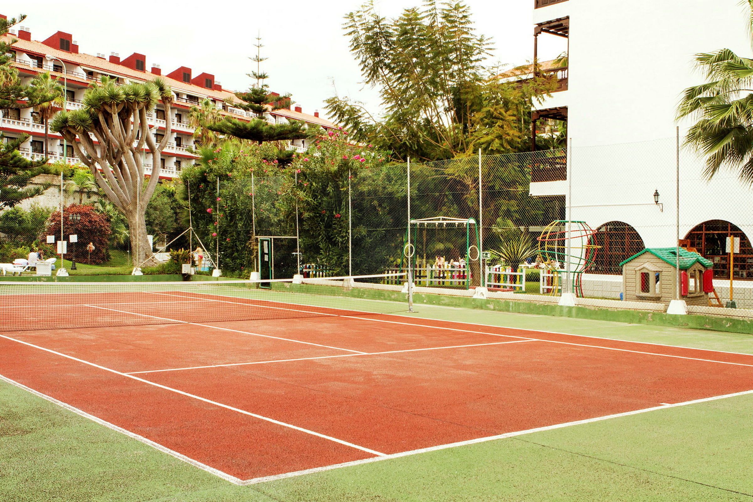 En tennisbane omgitt av planter og bygninger. (AI-generert tekst)