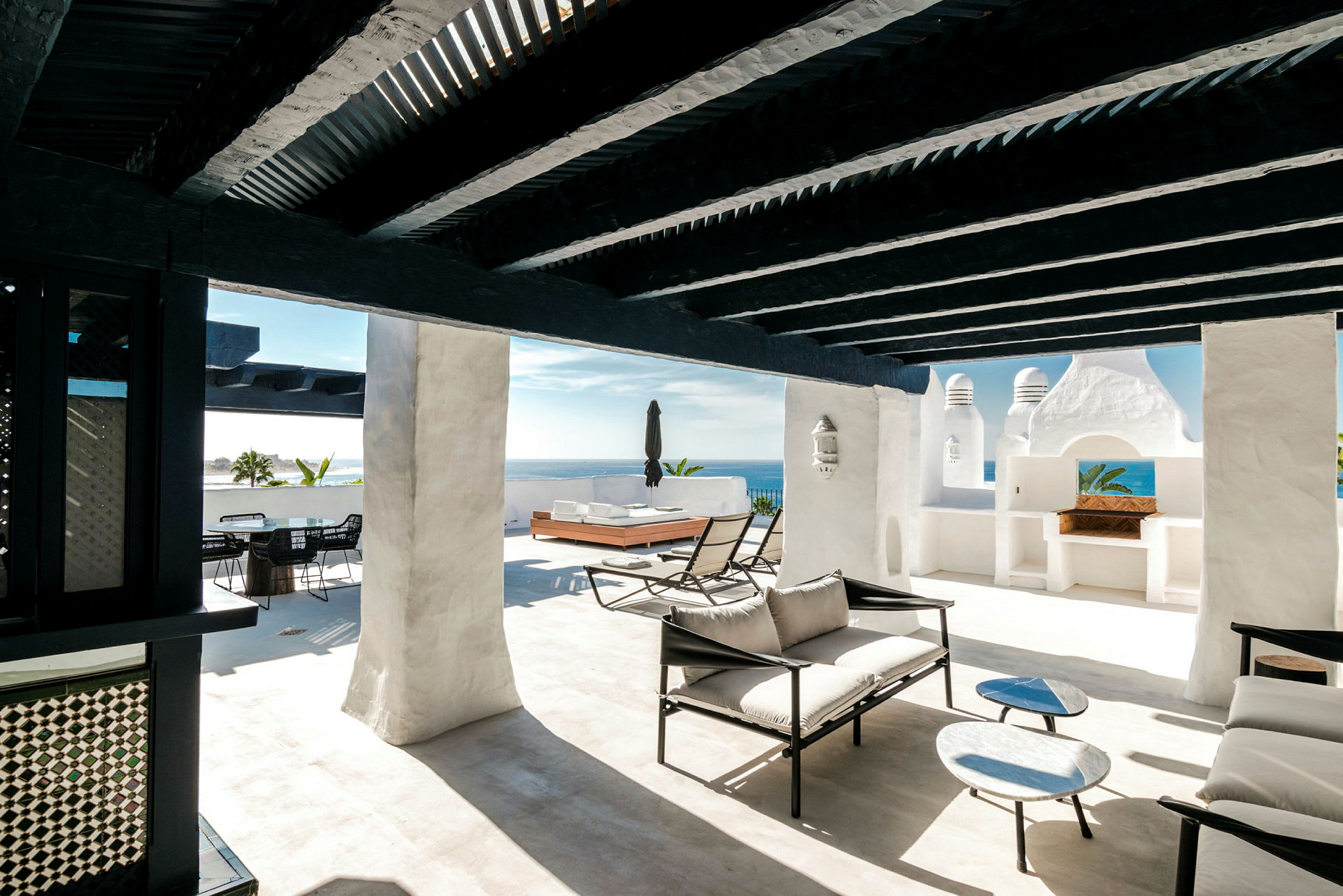 Solrik terrasse med hvite vegger, svarte trebjelker og moderne lounge møbler. (AI-generert tekst)