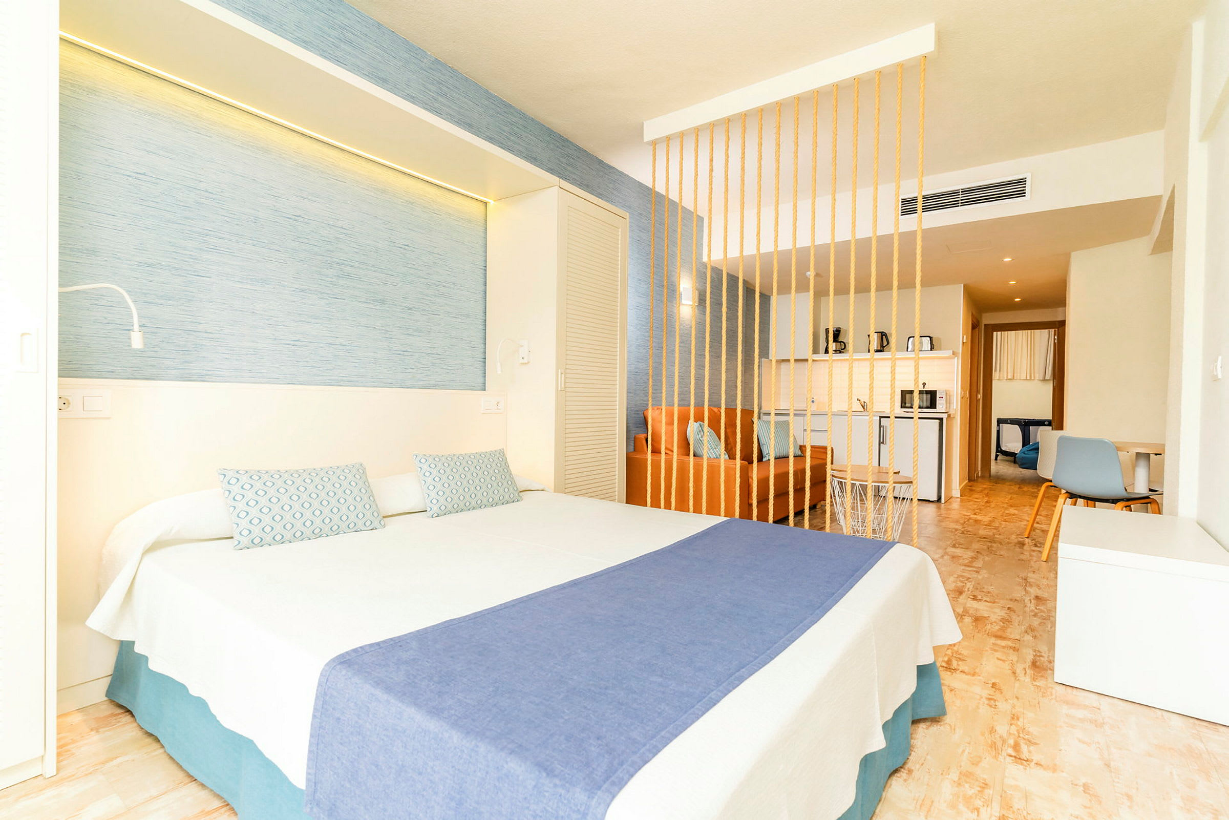 Hotellrom med en dobbeltseng, blått sengeteppe og dekorative puter, samt en oransje sofa bak en dekorativ skillevegg. (AI-generert tekst)