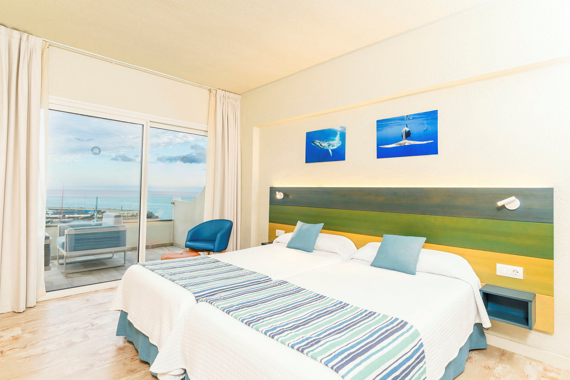 Hotellrom med to enkeltsenger, en grønn og blå sengegavl, samt en balkong med utsikt over havet. (AI-generert tekst)