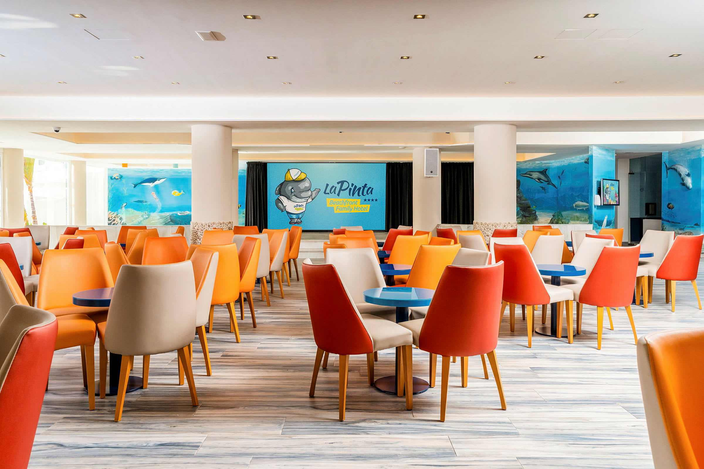 Spiseområde med fargerike stoler og bord, veggmaleri med tema fra havet inkludert delfiner og en tegnet figur som representerer hotellet. (AI-generert tekst)