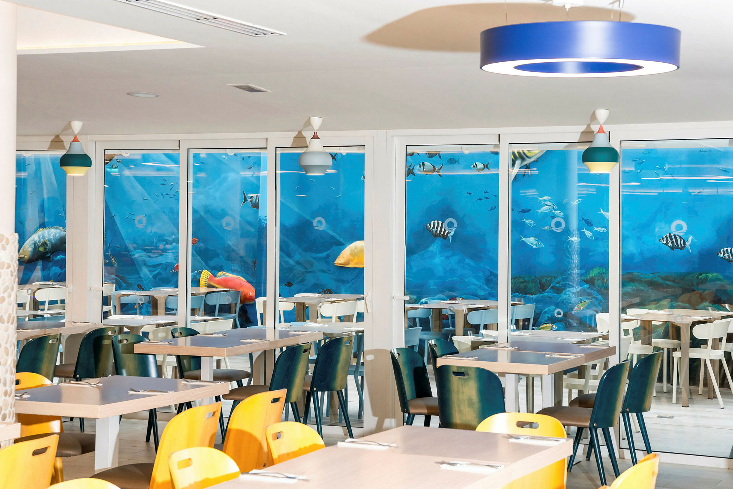 Restaurant med bord og stoler, med en vegg dekket av et blått akvariemotiv. (AI-generert tekst)