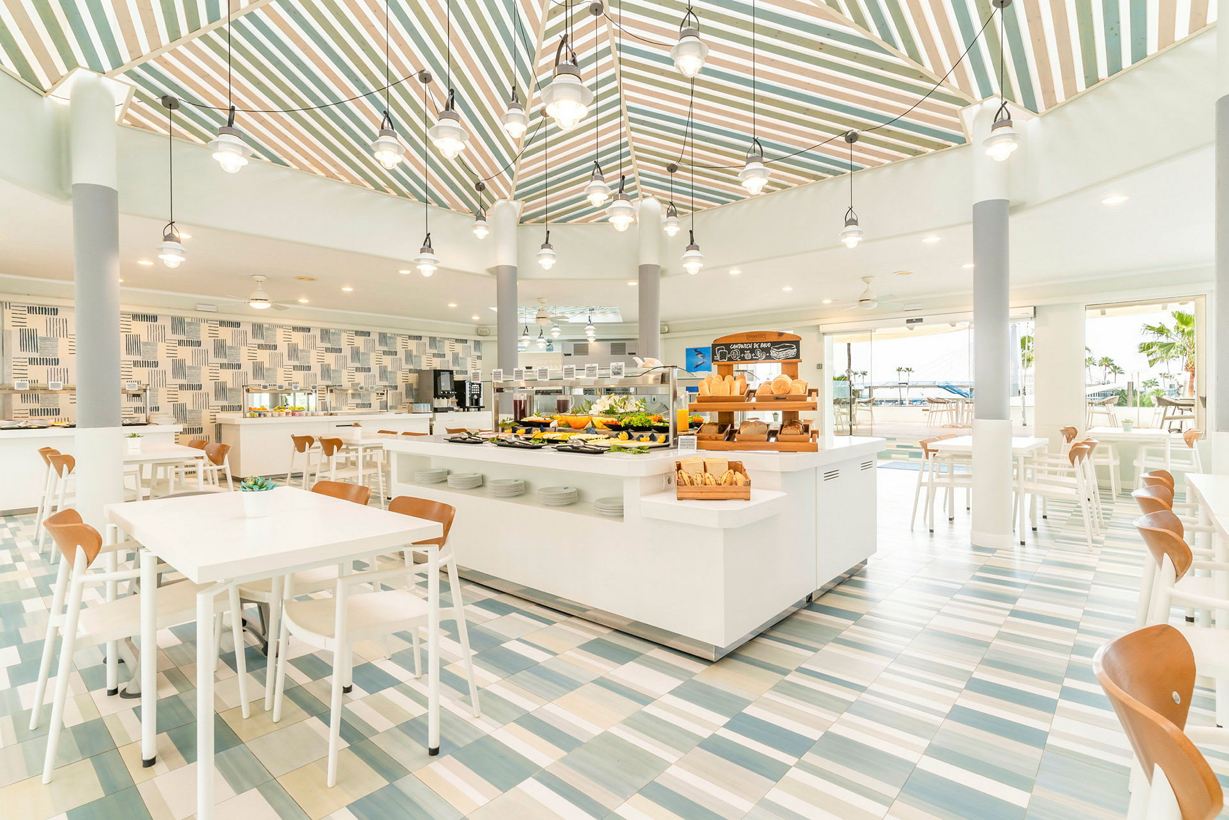 En lys og moderne restaurant med hvite møbler og fargerike fliser på gulvet. Et buffébord er sentralt plassert med forskjellige retter. (AI-generert tekst)