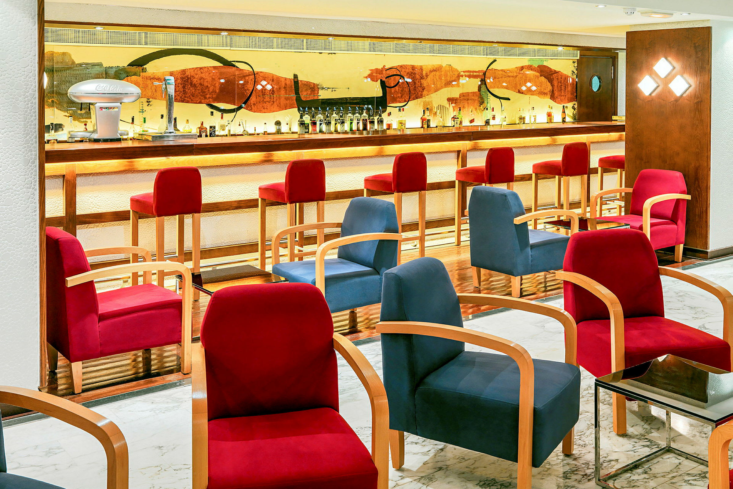 En hotellbar med en opplyst bardisk, høye røde stoler og komfortable sittemøbler i forskjellige farger. (AI-generert tekst)