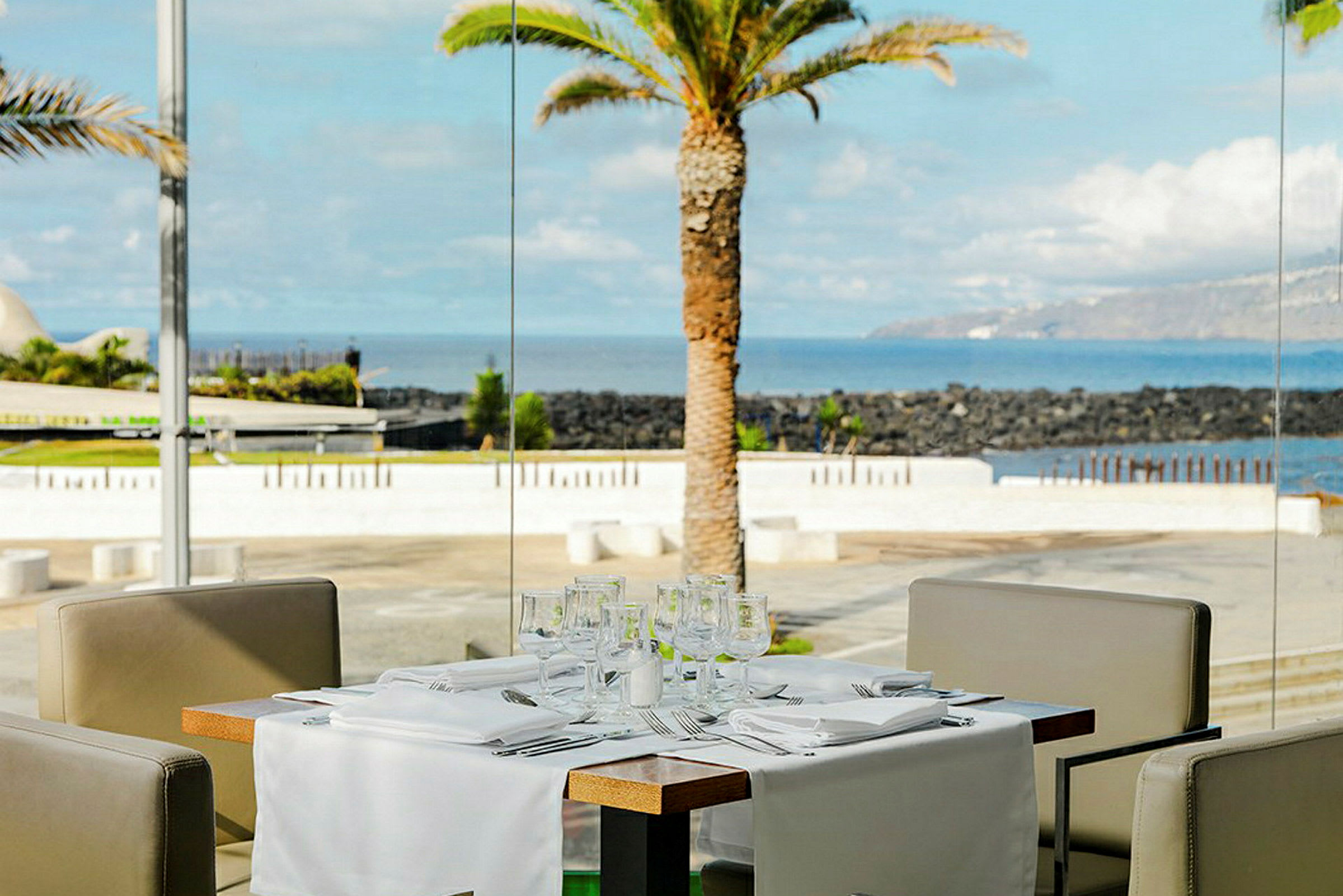 Et dekket utendørsbord i en restaurantmiljø med havutsikt og en palme i bakgrunnen. (AI-generert tekst)