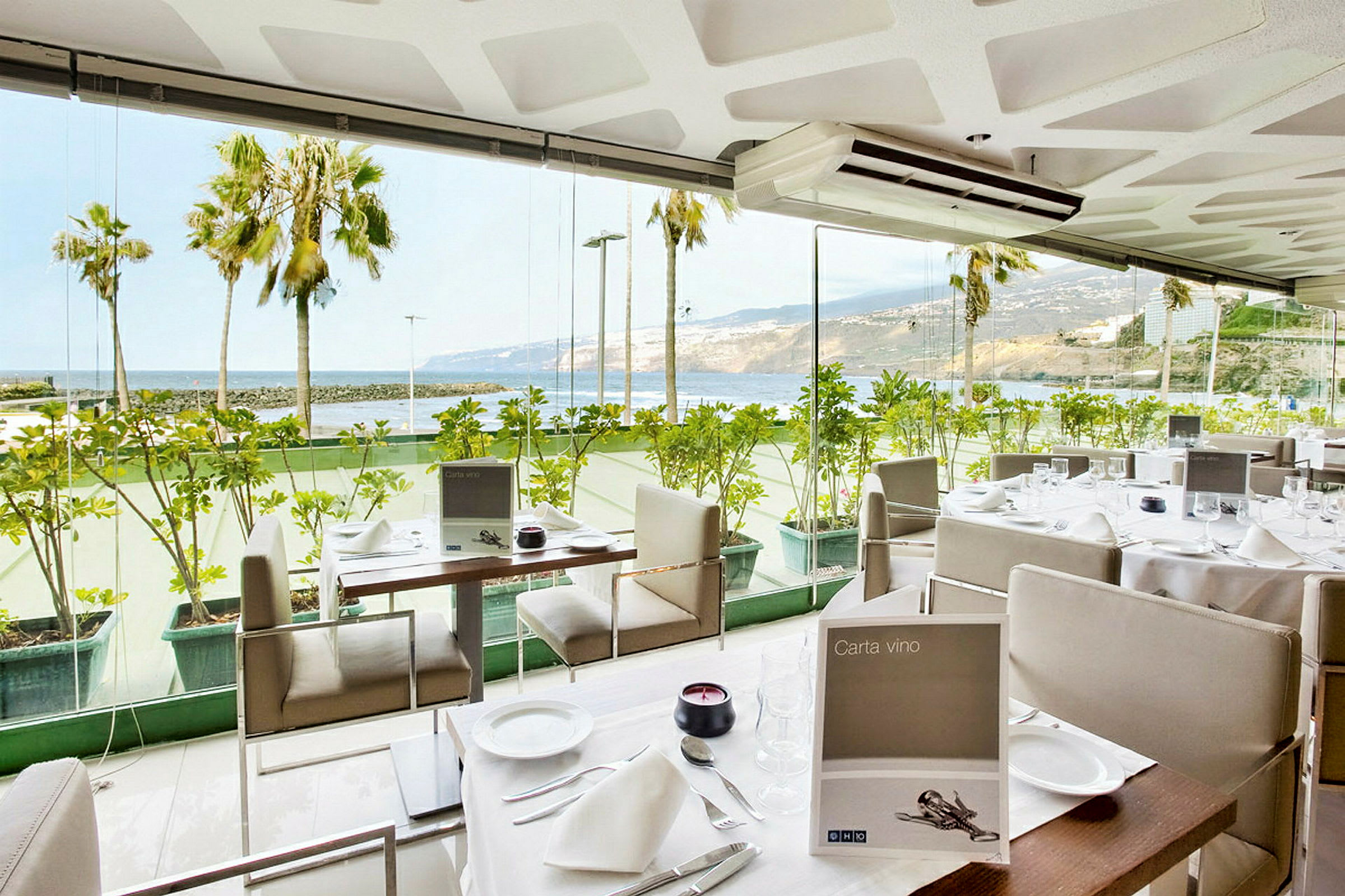 En restaurant med glassvegger og utsikt mot havet og palmer. (AI-generert tekst)
