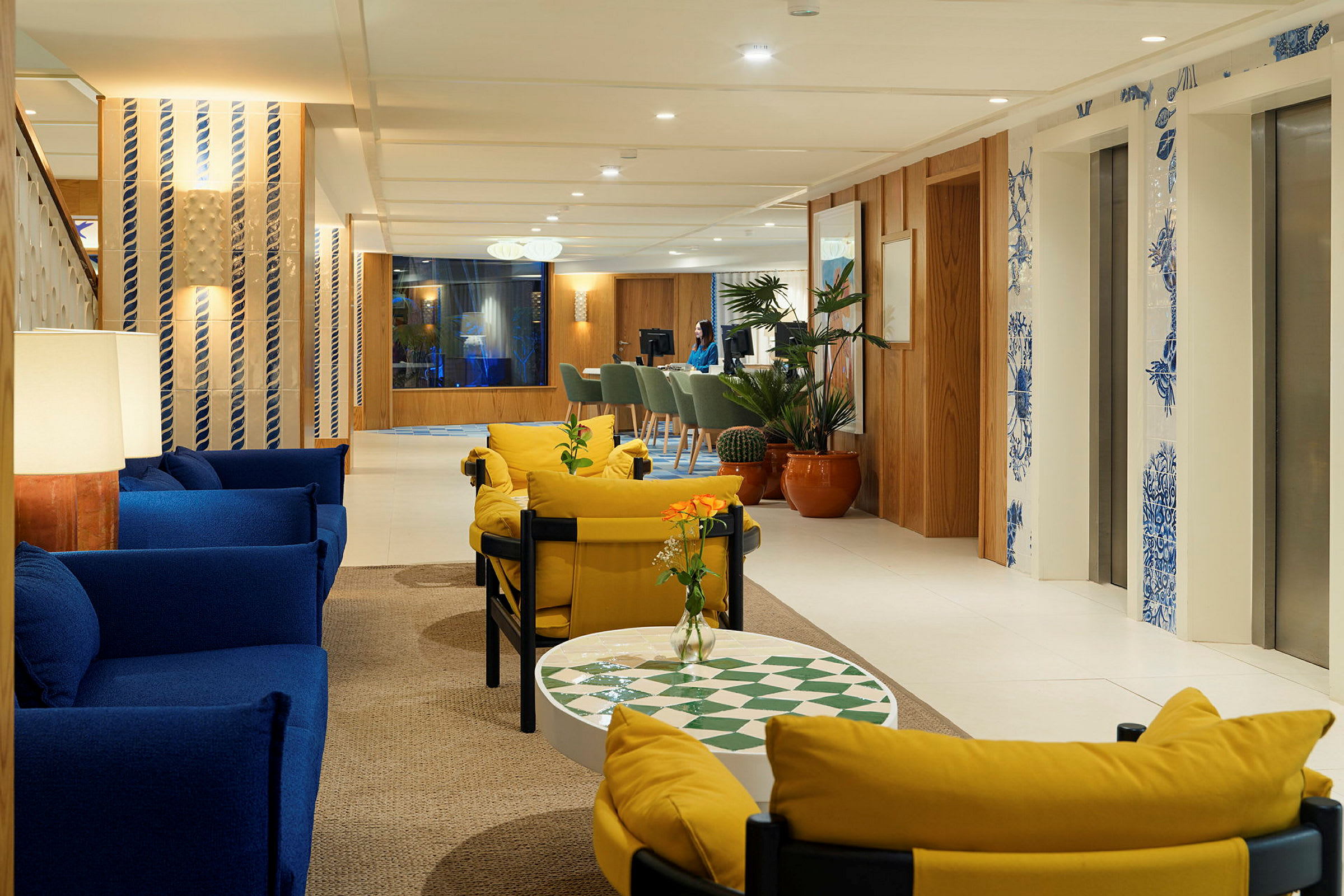 En lys hotellobby med sofaer i blått og gult, planter og heiser dekorert med blå detaljer. (AI-generert tekst)