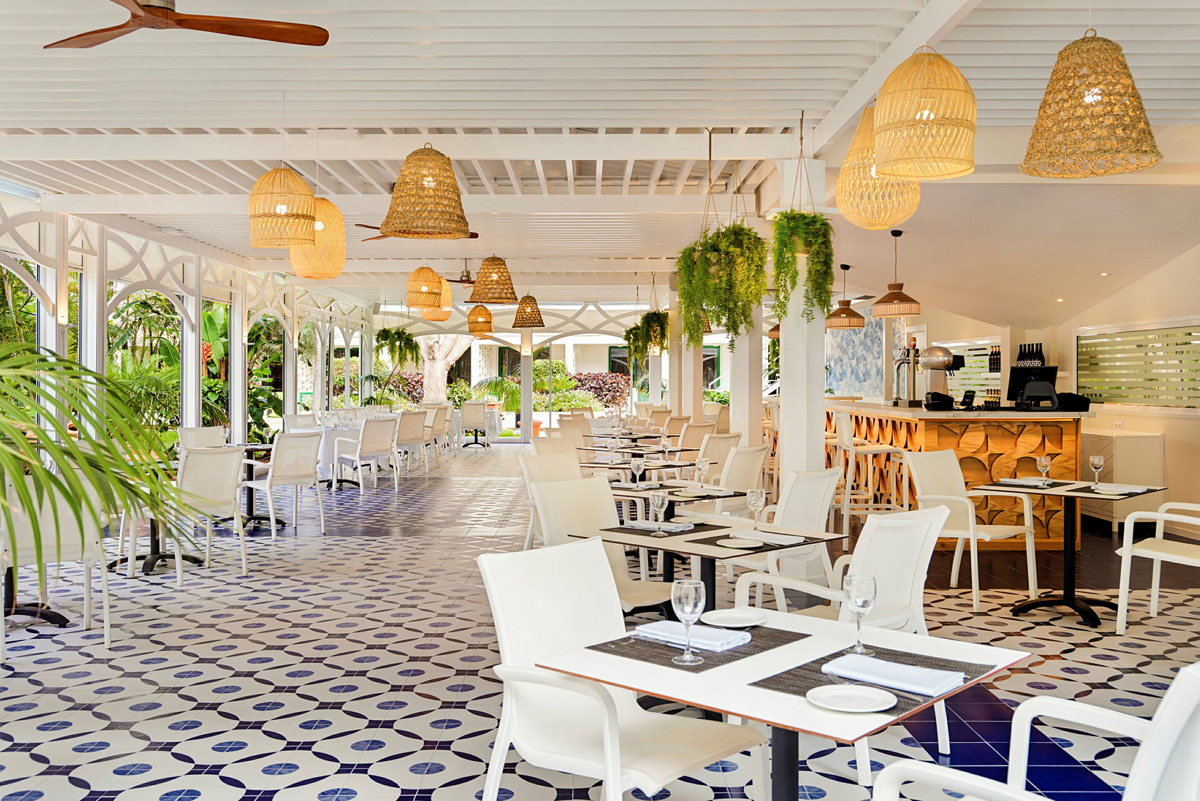 Bilde av en restaurant med hvite møbler, dekorativt flislagt gulv og taklamper i rotting. Grønne planter henger fra taket. (AI-generert tekst)