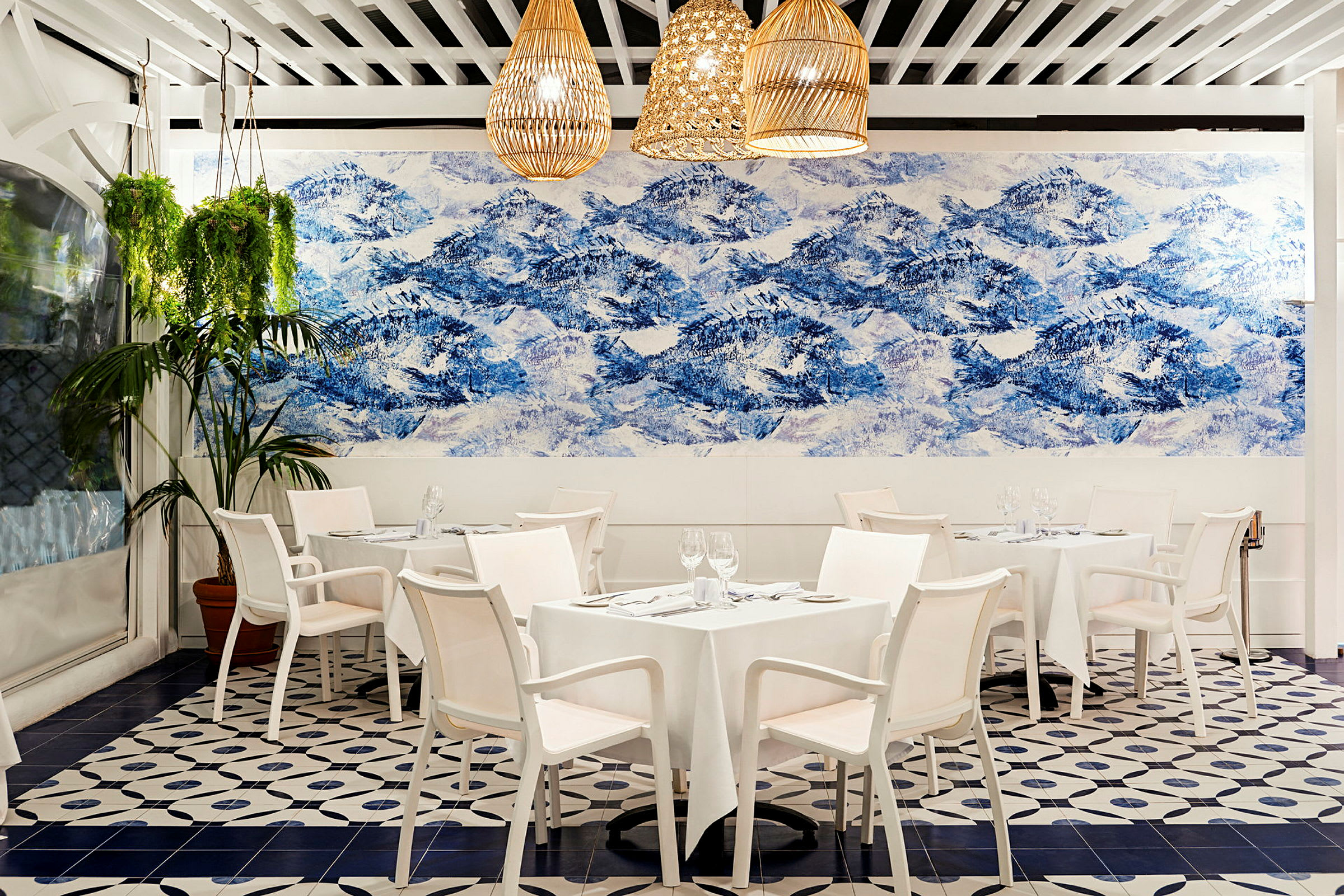 En innendørs restaurant med hvite bord og stoler, dekorert med et stort veggmaleri i blått og hvitt som forestiller fjell. Hengende planter pryder lokalet. (AI-generert tekst)