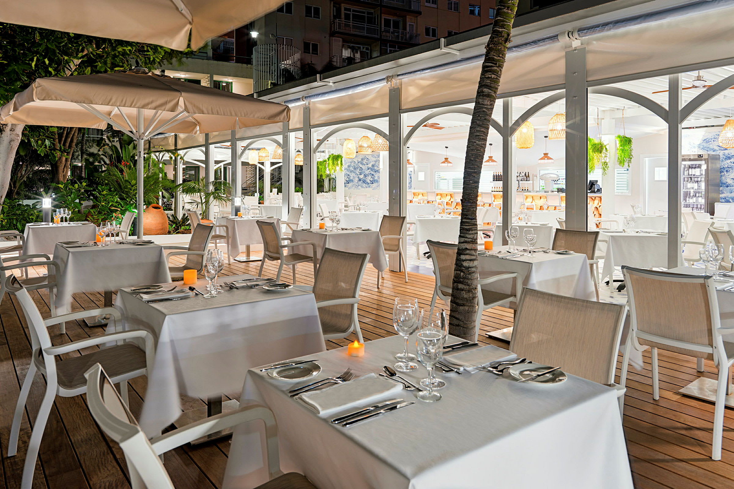 En utendørs restaurant med dekket bord og stoler, omgitt av grøntområder og opplyst av mykt lys. (AI-generert tekst)