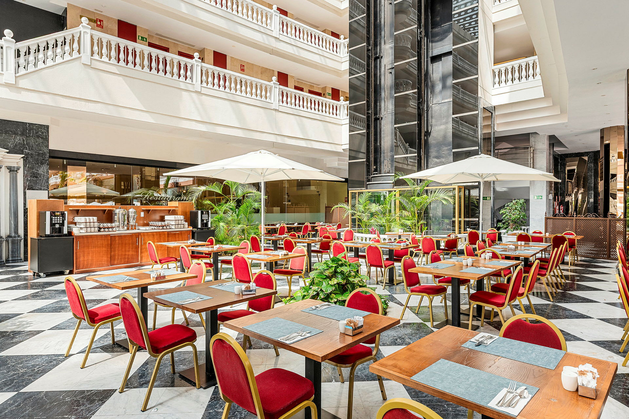 En restaurant med bord, røde stoler og parasoller i et lyst atrium. (AI-generert tekst)