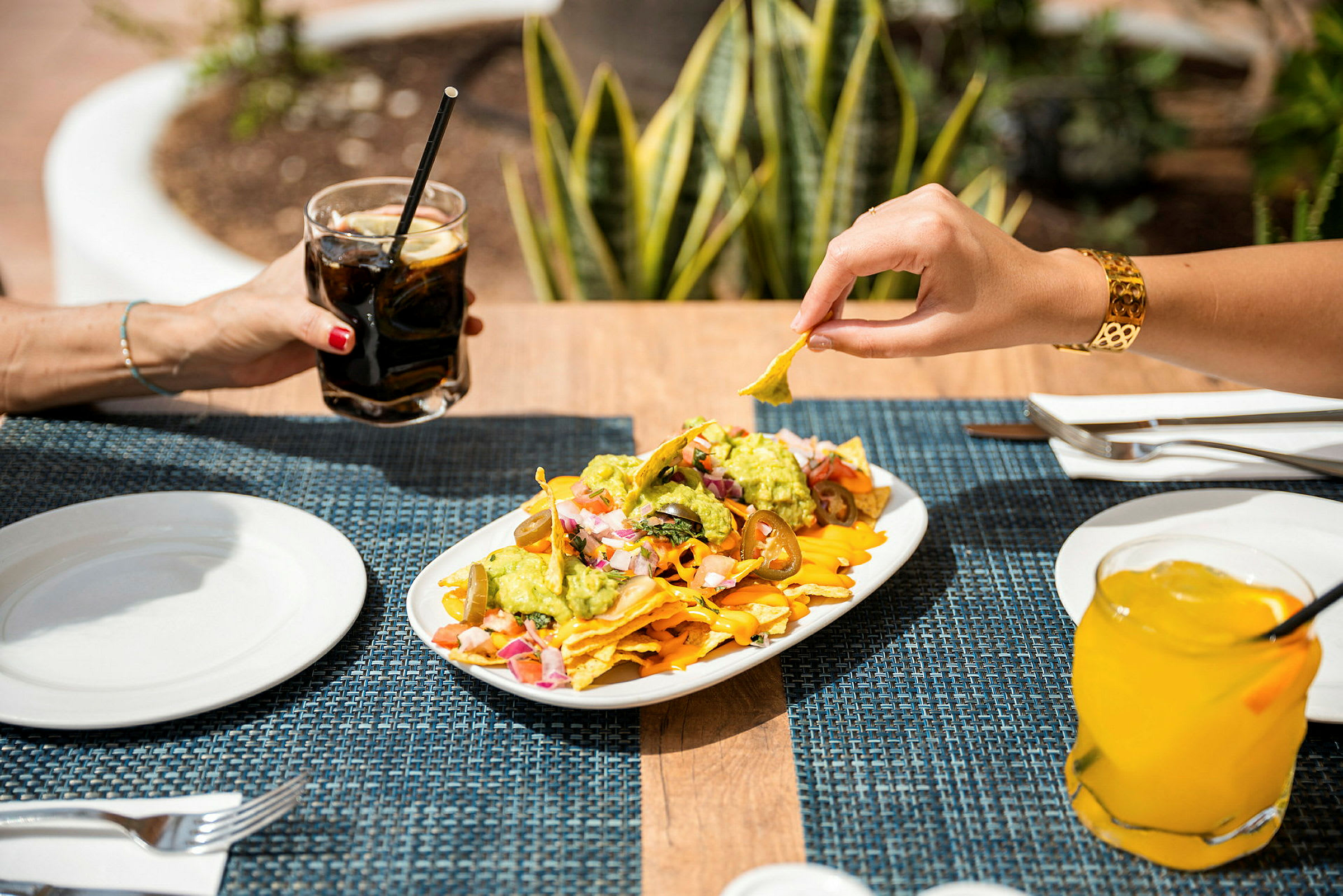 Tallerken med nachos, guacamole og tilbehør samt to glass med drikke på et dekket bord. (AI-generert tekst)