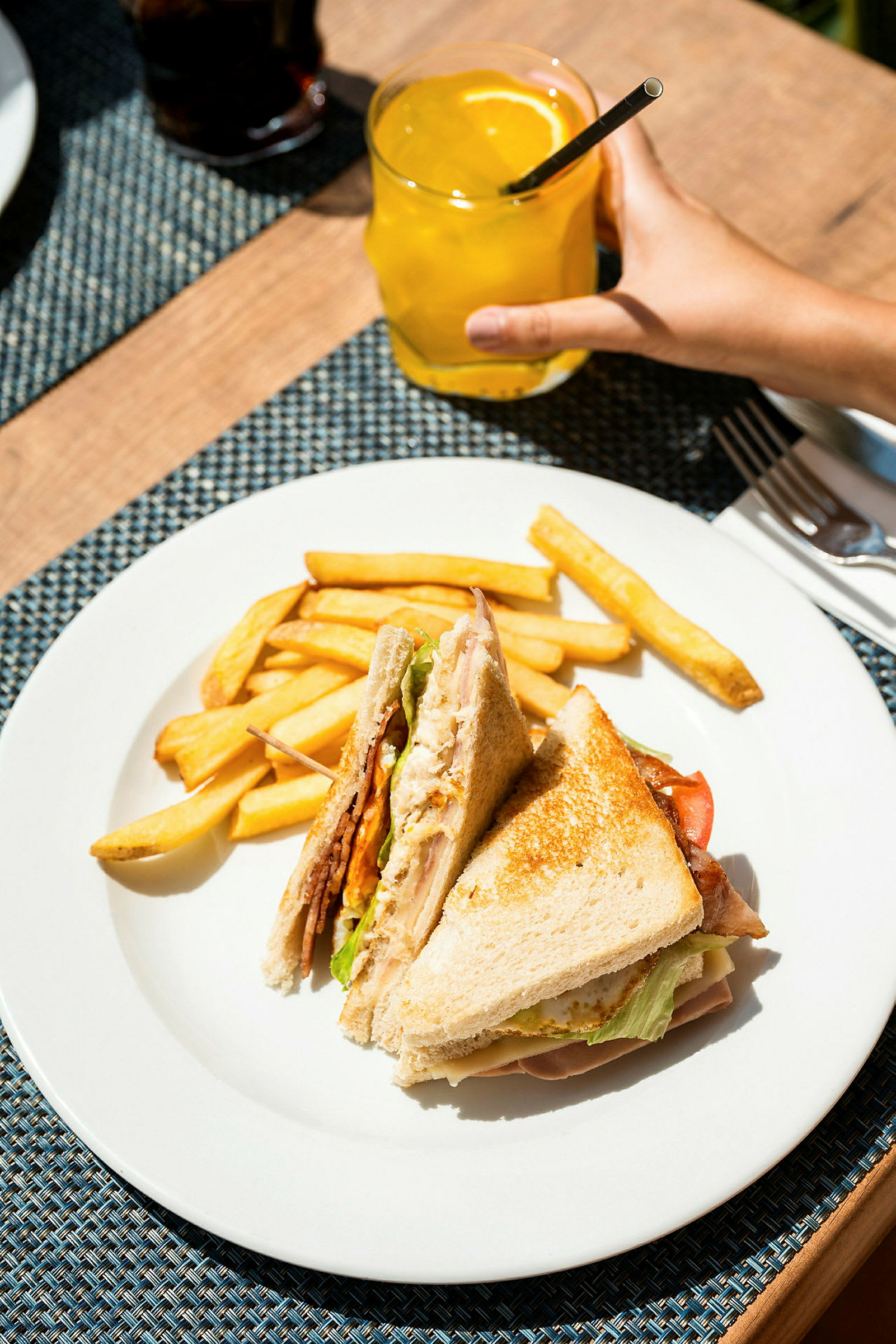 En tallerken med en sandwich og pommes frites, samt en hånd som holder et glass med en gul drikke. (AI-generert tekst)