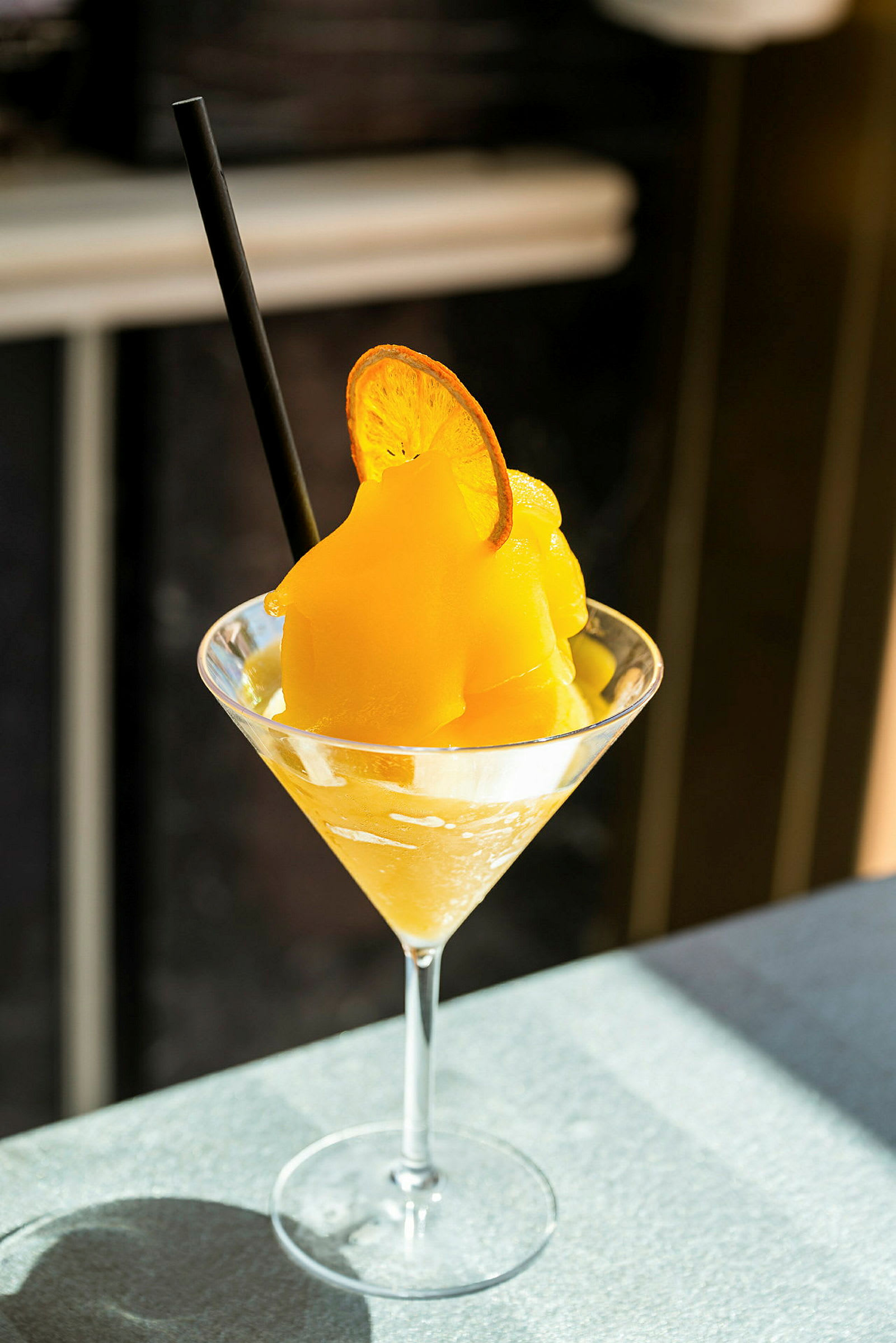 Et cocktailglass med frossen gul drikk toppet med en tørket appelsinskive og et svart sugerør. (AI-generert tekst)