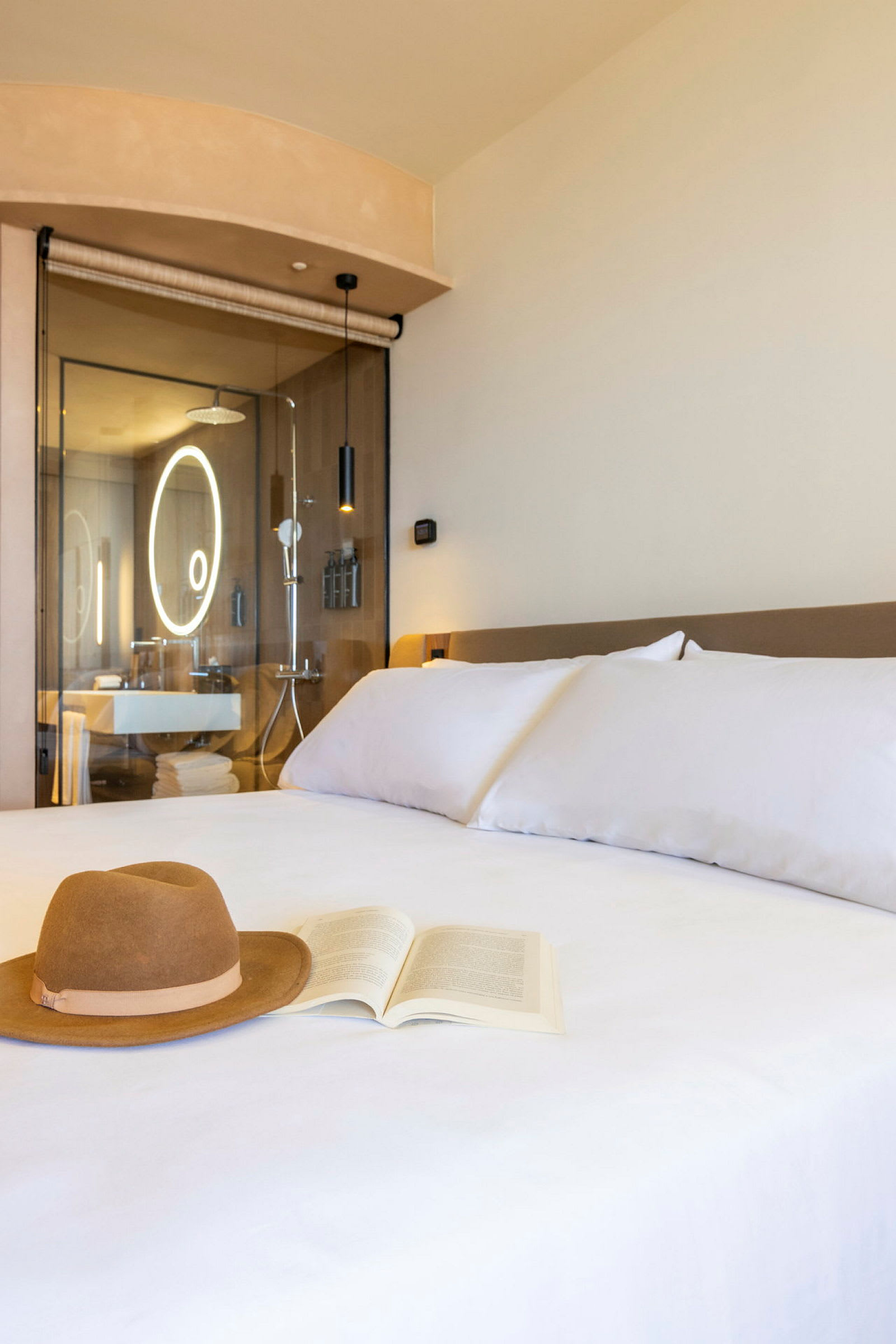 Et hotellrom med en seng der en hatt og en bok ligger. I bakgrunnen vises et moderne bad. (AI-generert tekst)
