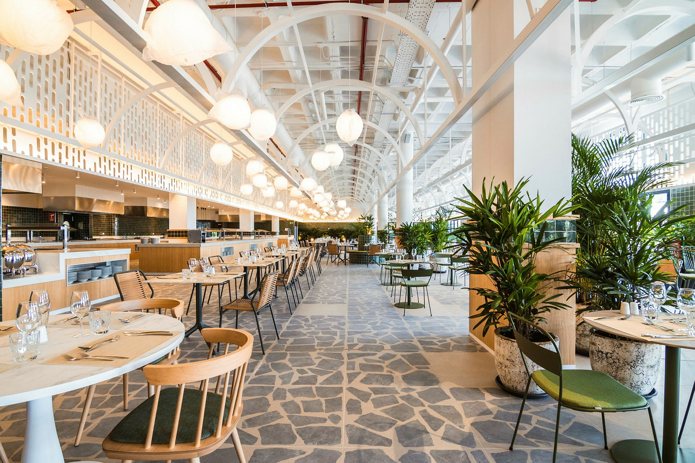 En moderne og lys restaurant med bord og stoler samt grønne planter. Takhøyde og belysning bidrar til en luftig atmosfære. (AI-generert tekst)