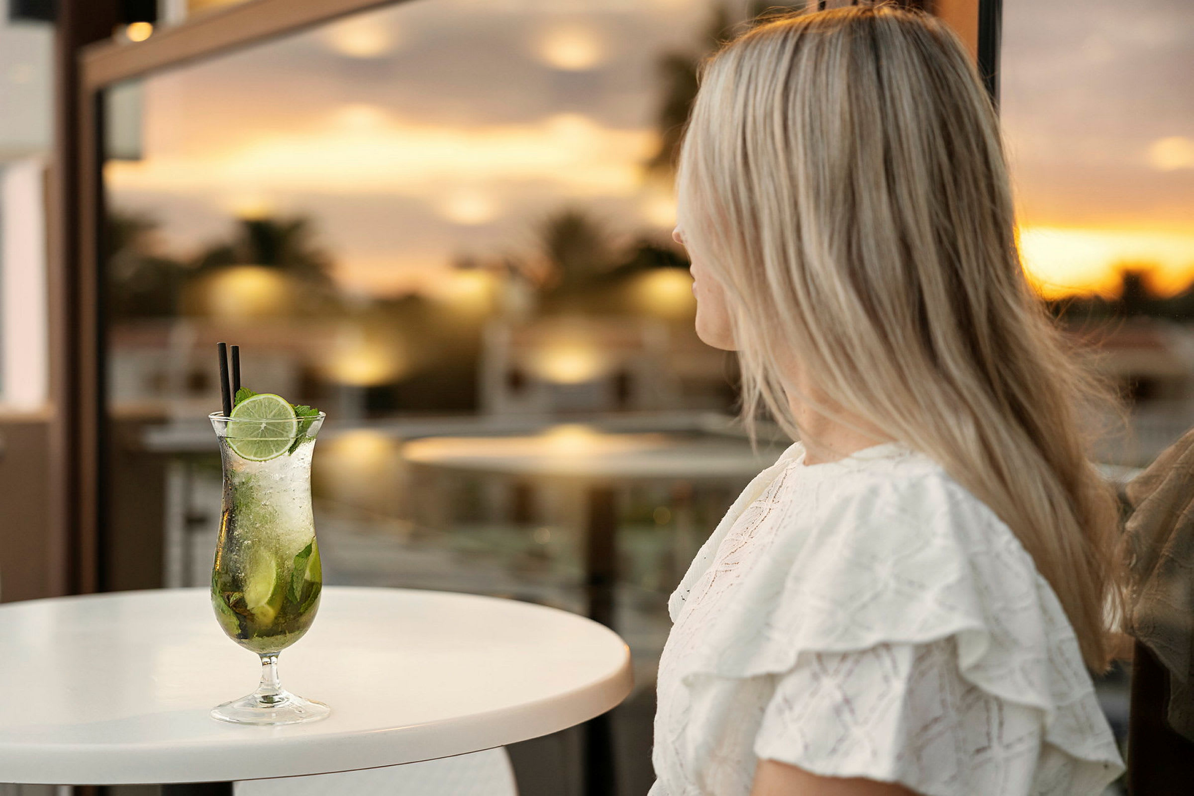 Kvinne med langt lyst hår sitter ved et bord med en cocktail foran seg, bakgrunn er en solnedgang. (AI-generert tekst)