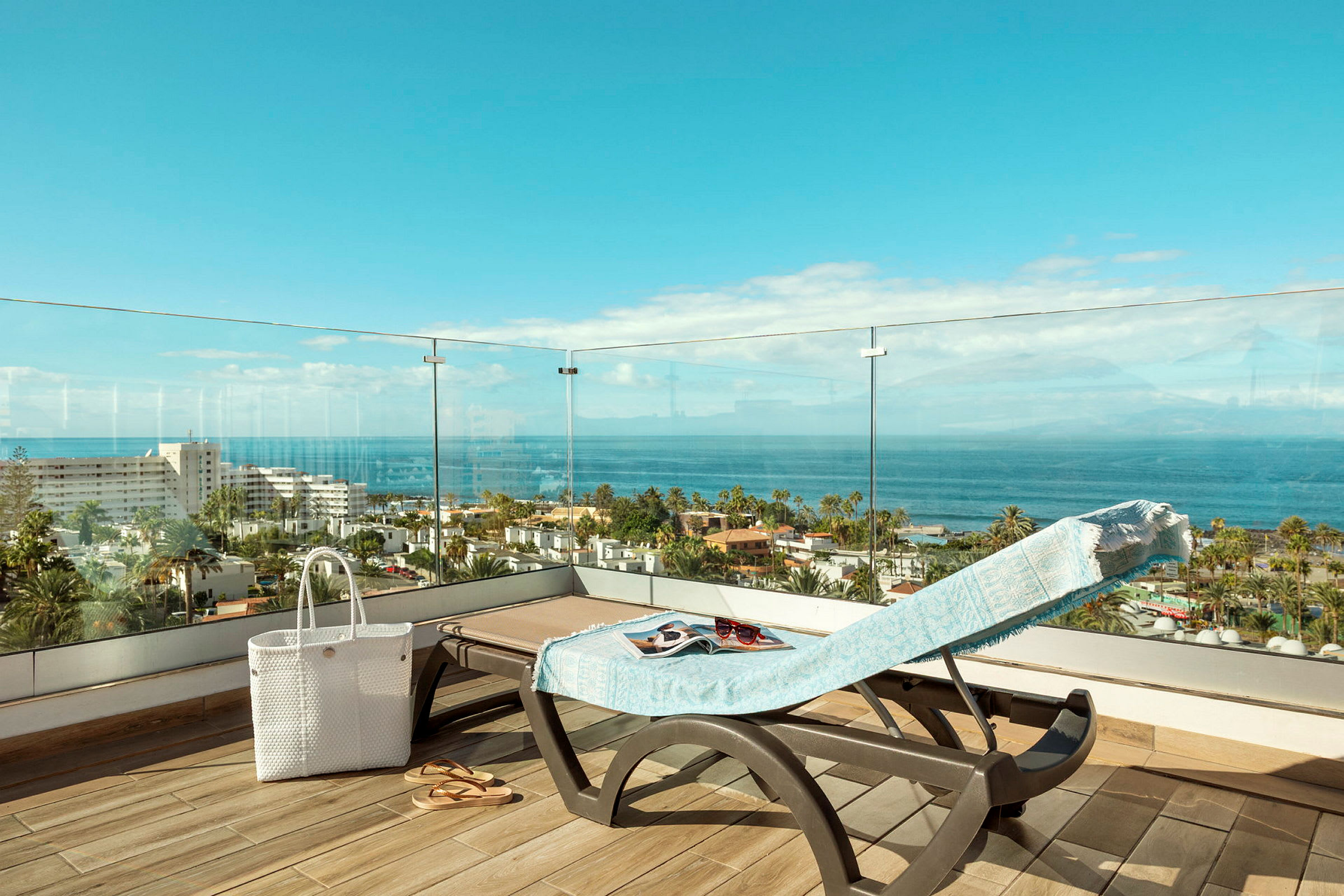 Utsikt fra en terrasse med en solseng dekket med et håndkle, en hvit veske og sandaler, over man ser havet og bygninger. (AI-generert tekst)