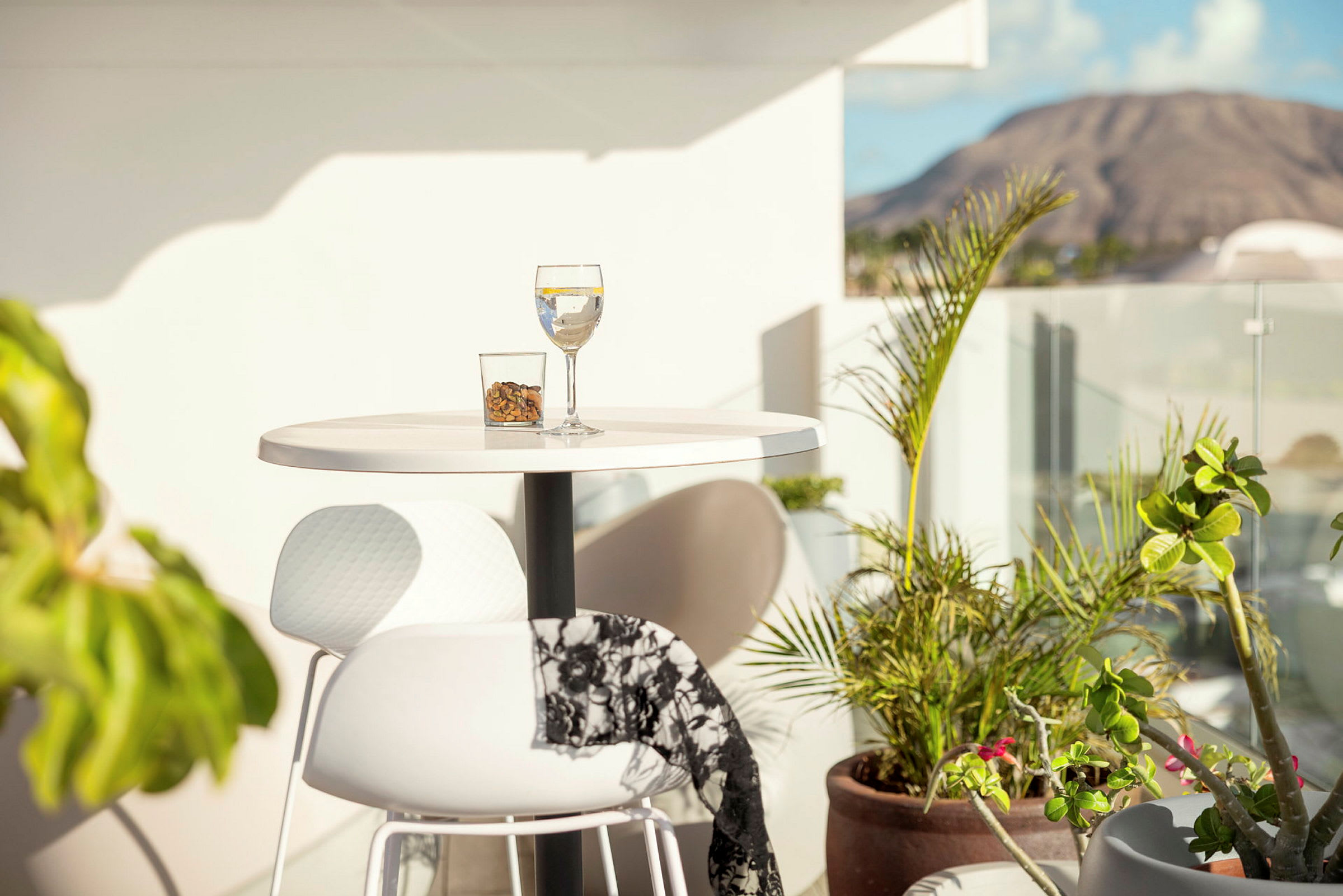 Et lite bord på en terrasse med et glass drikke og en skål med nøtter i solen, omgitt av planter og utsikt mot et fjell. (AI-generert tekst)