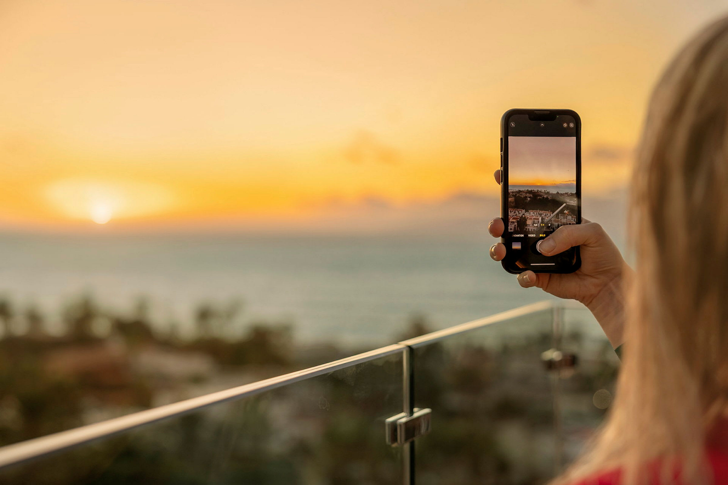 Person tar et bilde med mobilkameraet sitt av en solnedgang over havet fra en balkong. (AI-generert tekst)