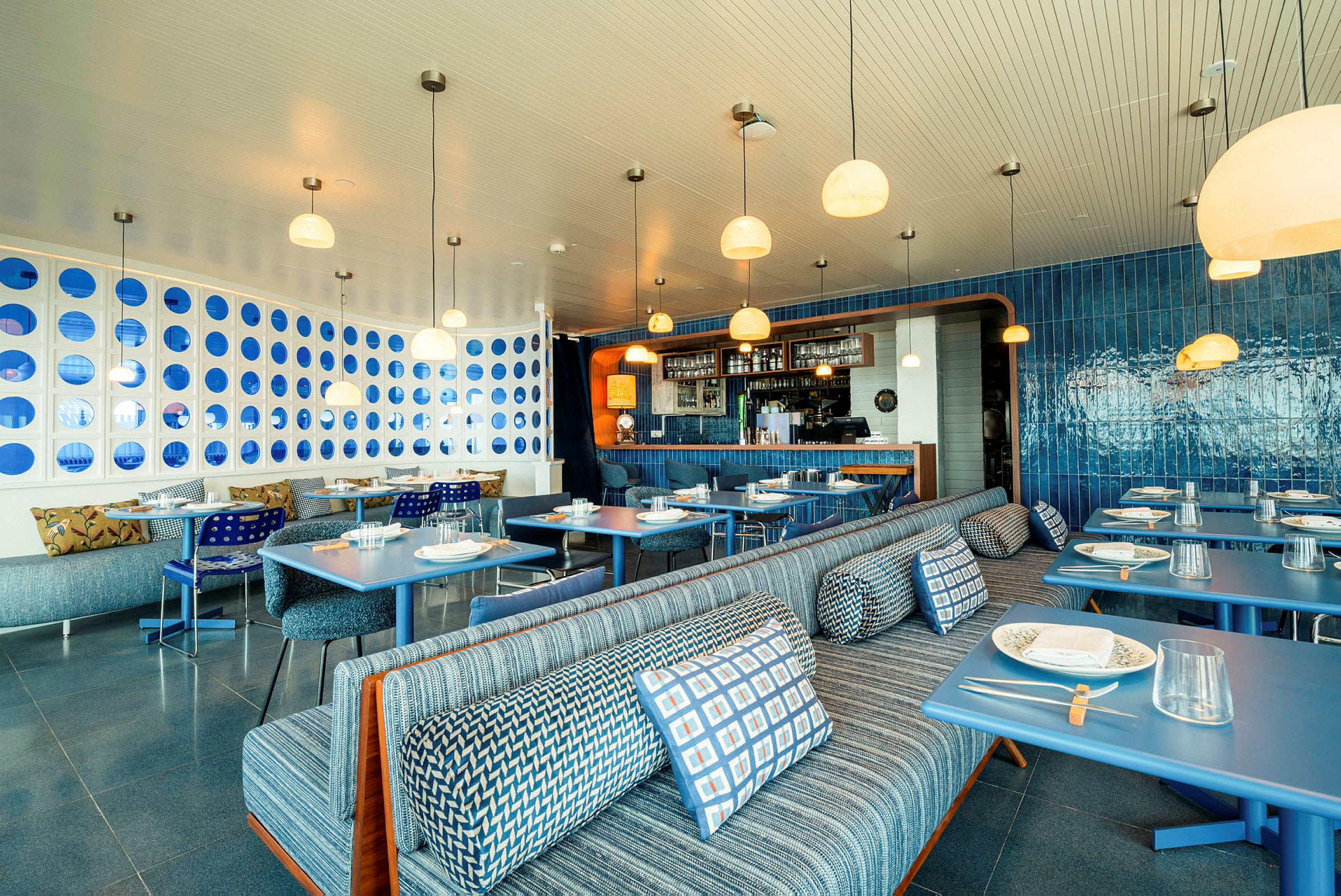 Bilde av en moderne restaurant med elegant interiør i blåtoner, sitteplasser med mønstrede puter og dekkede bord. (AI-generert tekst)